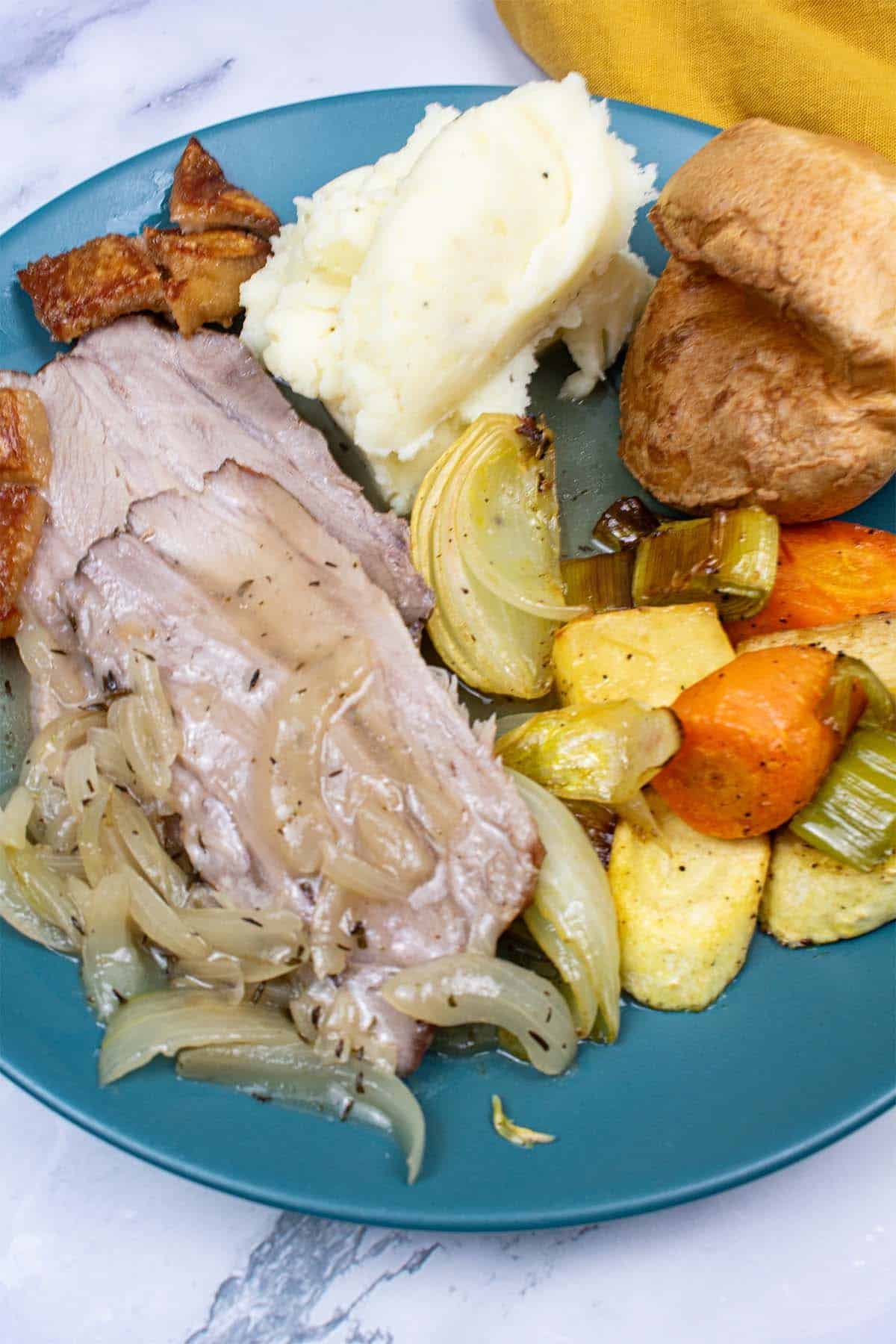 Roast pork with crackling, roasted root vegetables, mashed potatoes, a yorkshire pudding plus cider and onion gravy on a teal dinner plate. Mustard napkin in the background.