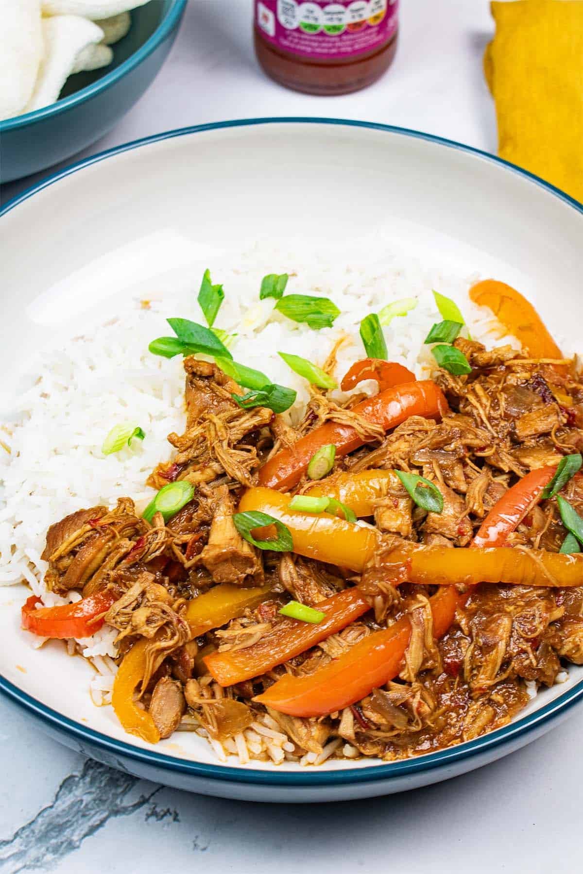 Sweet and sour chicken in a teal rimmed bowl with rice, a bowl of prawn crackers, bottle of sweet chilli sauce and mustard napkin in the background.