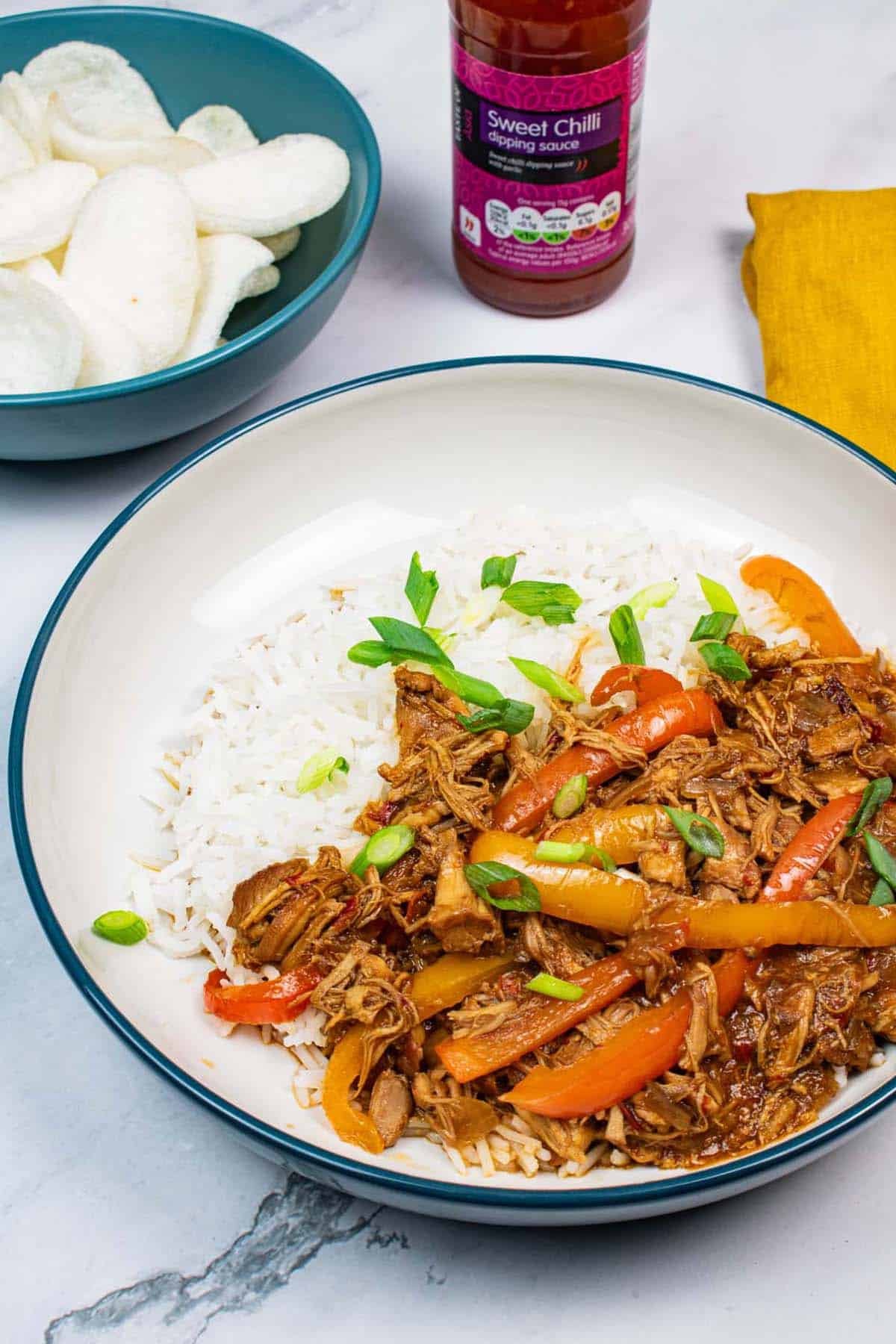 Sweet and sour chicken in a teal rimmed bowl with rice, a bowl of prawn crackers, bottle of sweet chilli sauce and mustard napkin in the background.