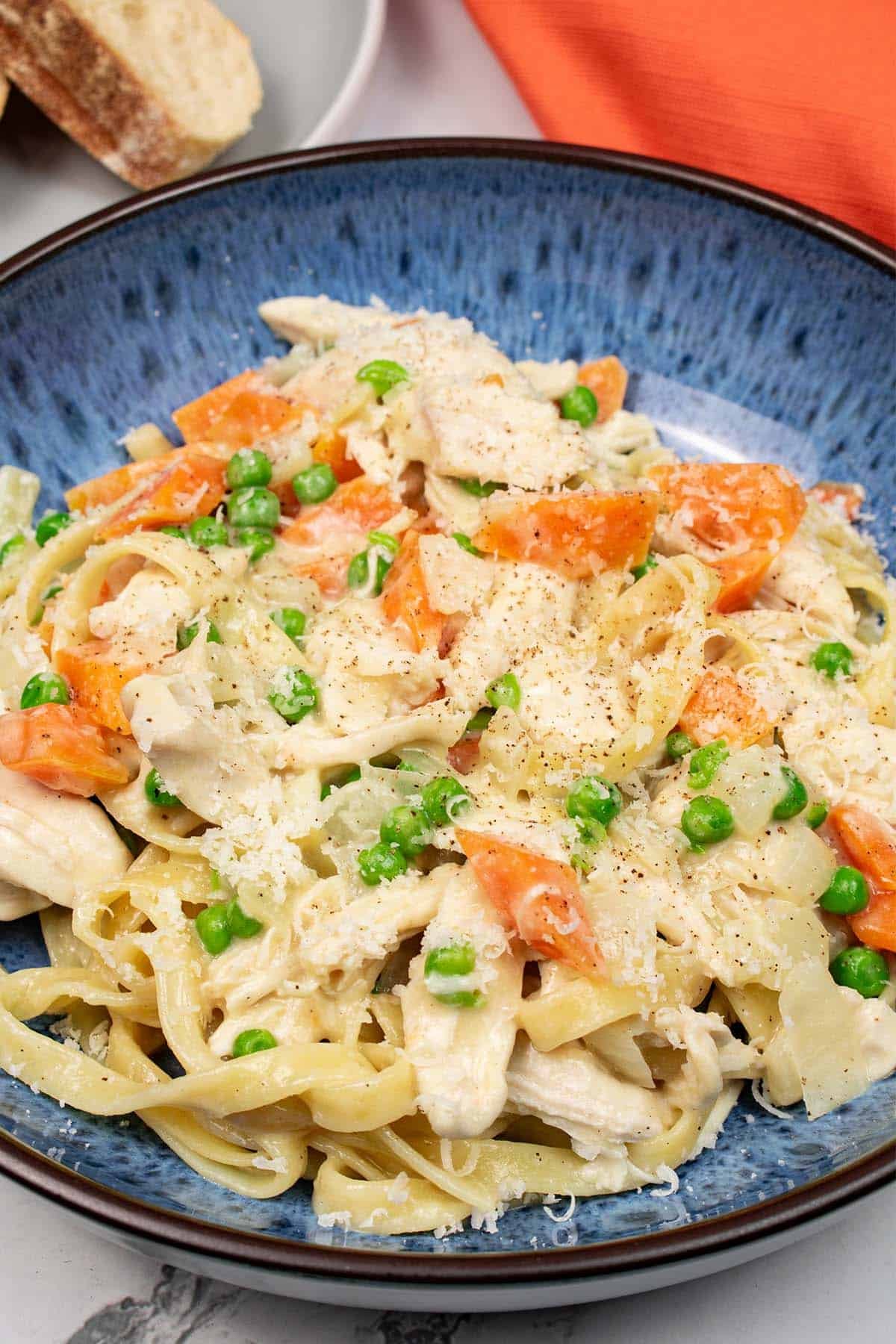 Creamy chicken and vegetable tagliatelle in a blue pattered bowl with slices of crusty bread on a grey side plate, and orange napkin on the side.