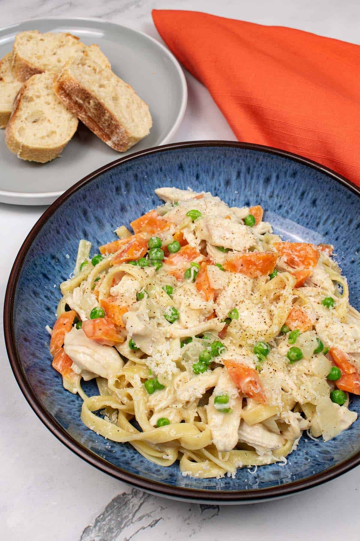 Creamy chicken and vegetable tagliatelle in a blue pattered bowl with slices of crusty bread on a grey side plate, and orange napkin on the side.