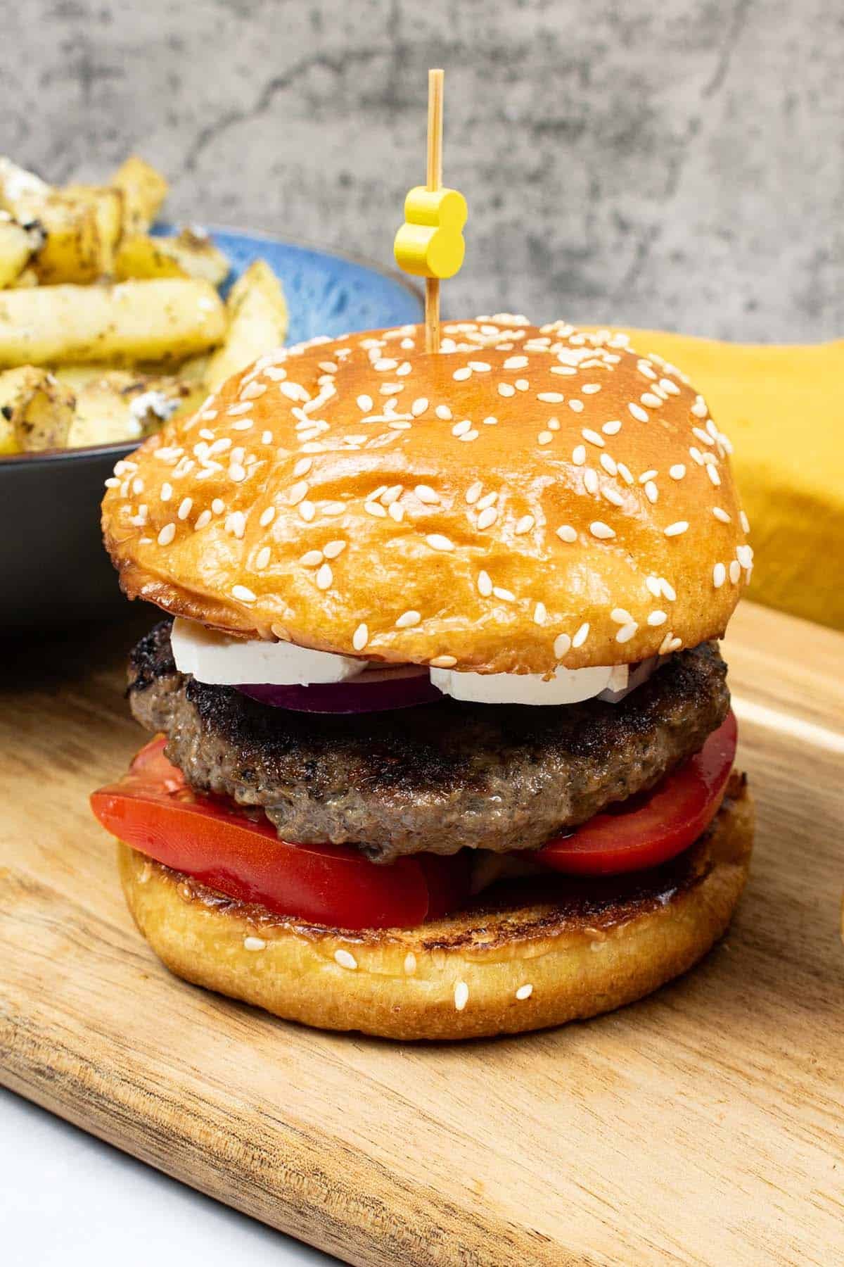 A lamb burger in a seeded brioche bun on a wooden board with a bowl of feta and oregano fries on the side.
