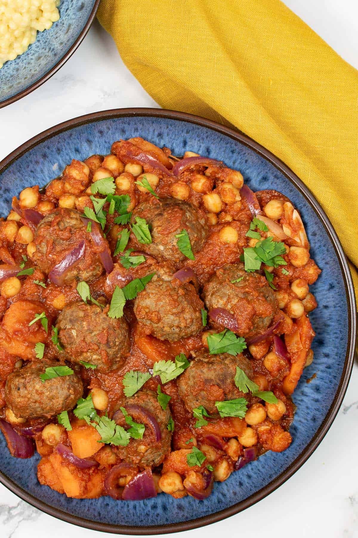 Lamb meatball tagine in a blue patterned bowl, with a bowl of couscous and mustard napkin on the side.