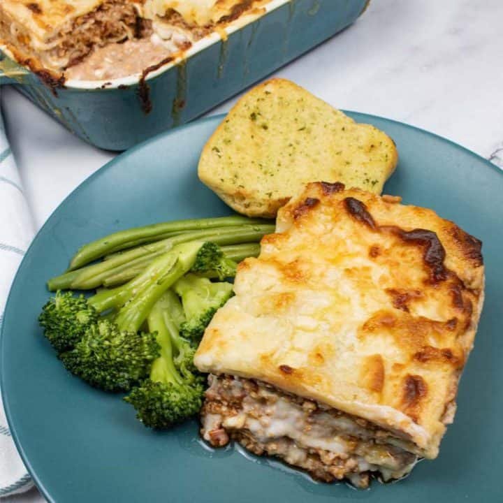 A portion of lasagne bolognese on a teal plate with broccoli and beans and a slice of garlic bread with the Staub dish and teal and white checked towel on the side.