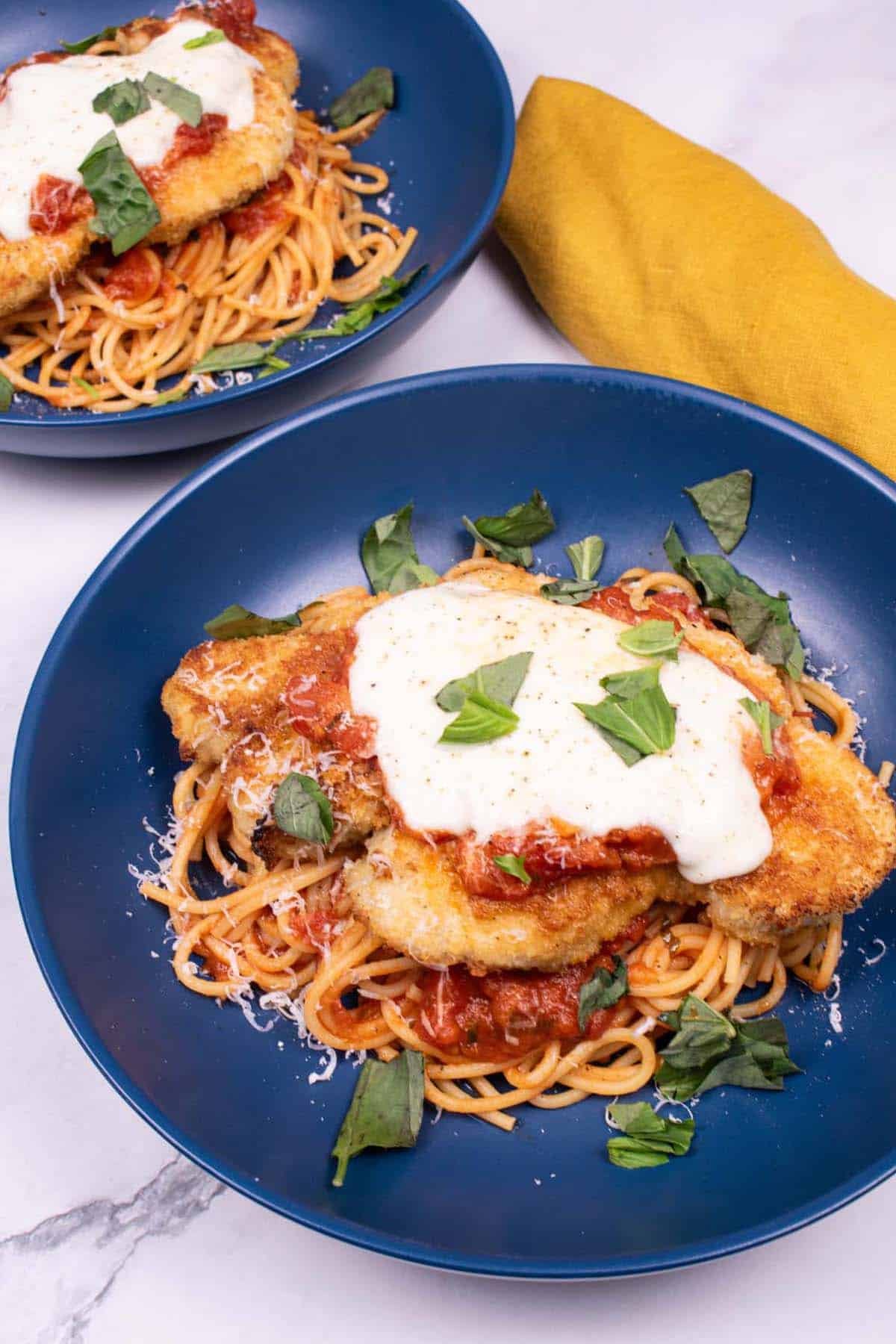 2 bowls of turkey parmigiana with spaghetti in blue bowls with a mustard napkin on the side.