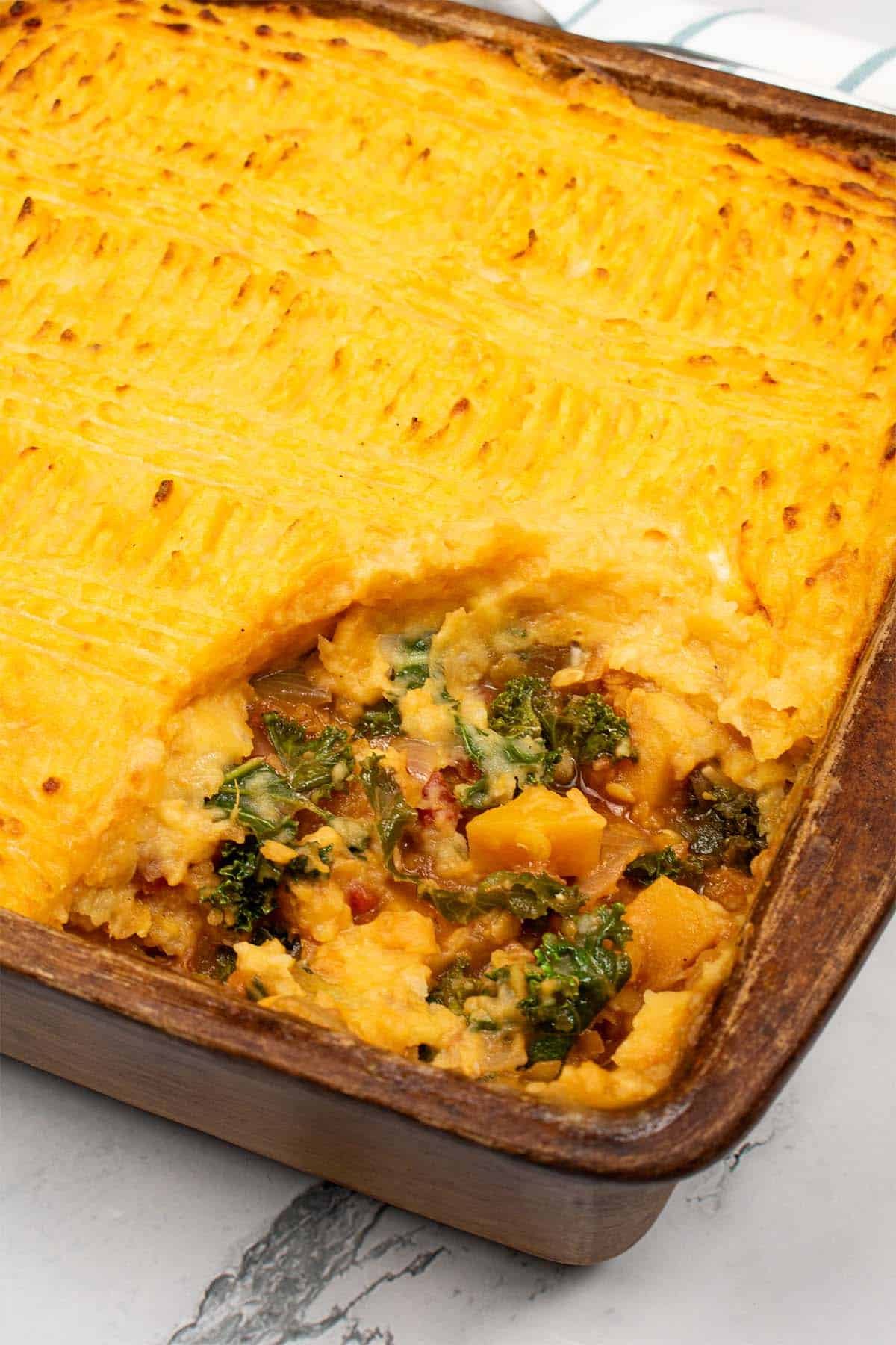 Vegetable and lentil cottage pie in a square stoneware dish with a serving spoon and teal and white towel on the side.