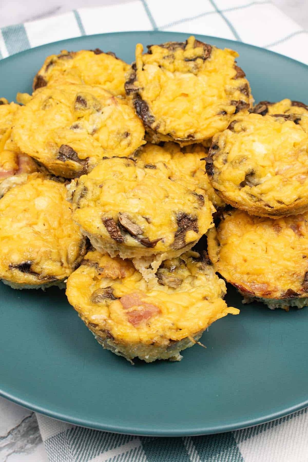12 cheese, ham and mushroom omelette muffins on a teal plate, on top of a teal and white checked towel.