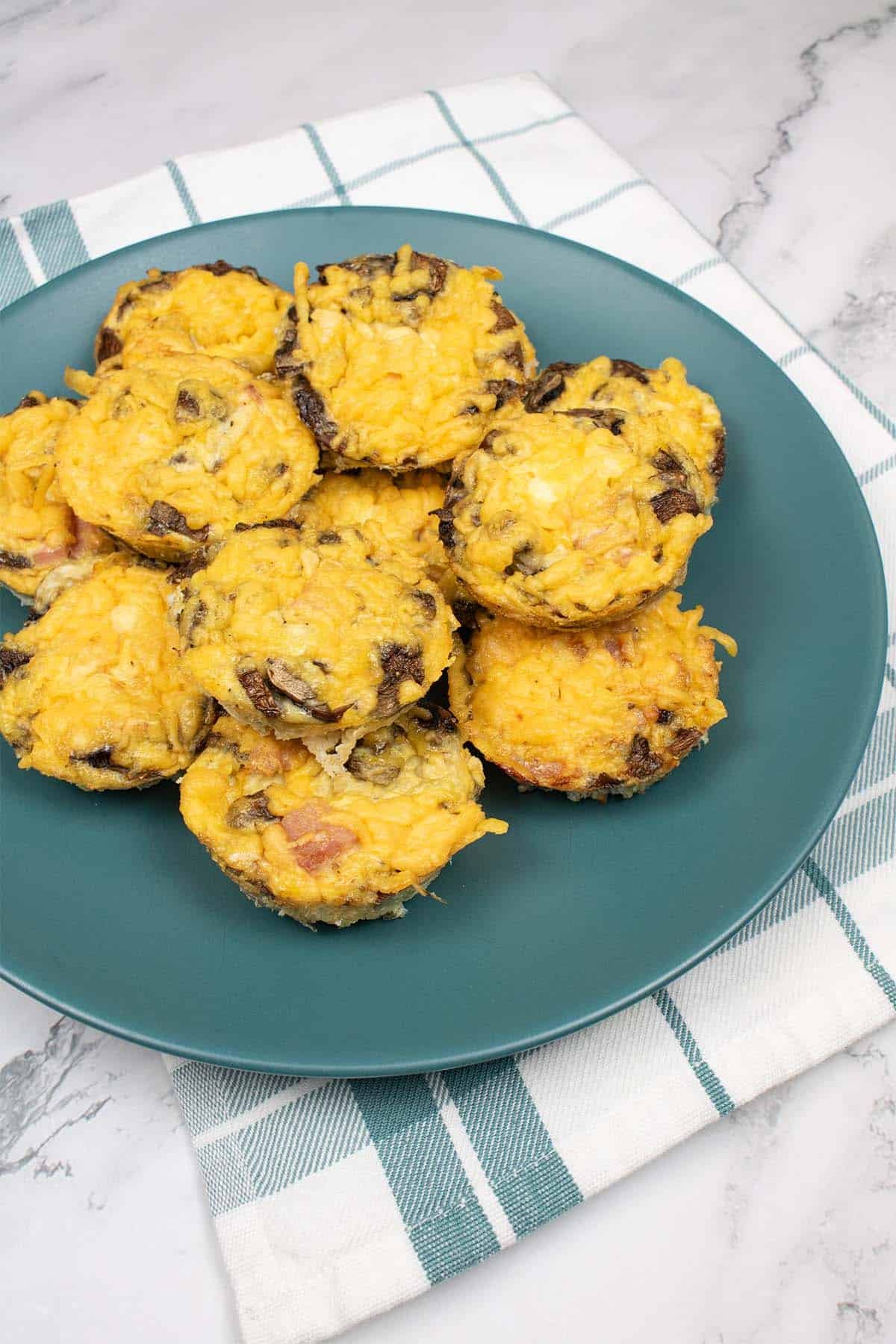 12 cheese, ham and mushroom omelette muffins on a teal plate, on top of a teal and white checked towel.