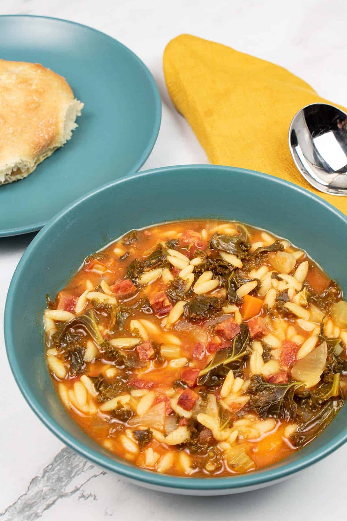 Chorizo and orzo soup in a teal bowl with a side plate and ciabatta roll, soup spoon and mustard napkin on the side.