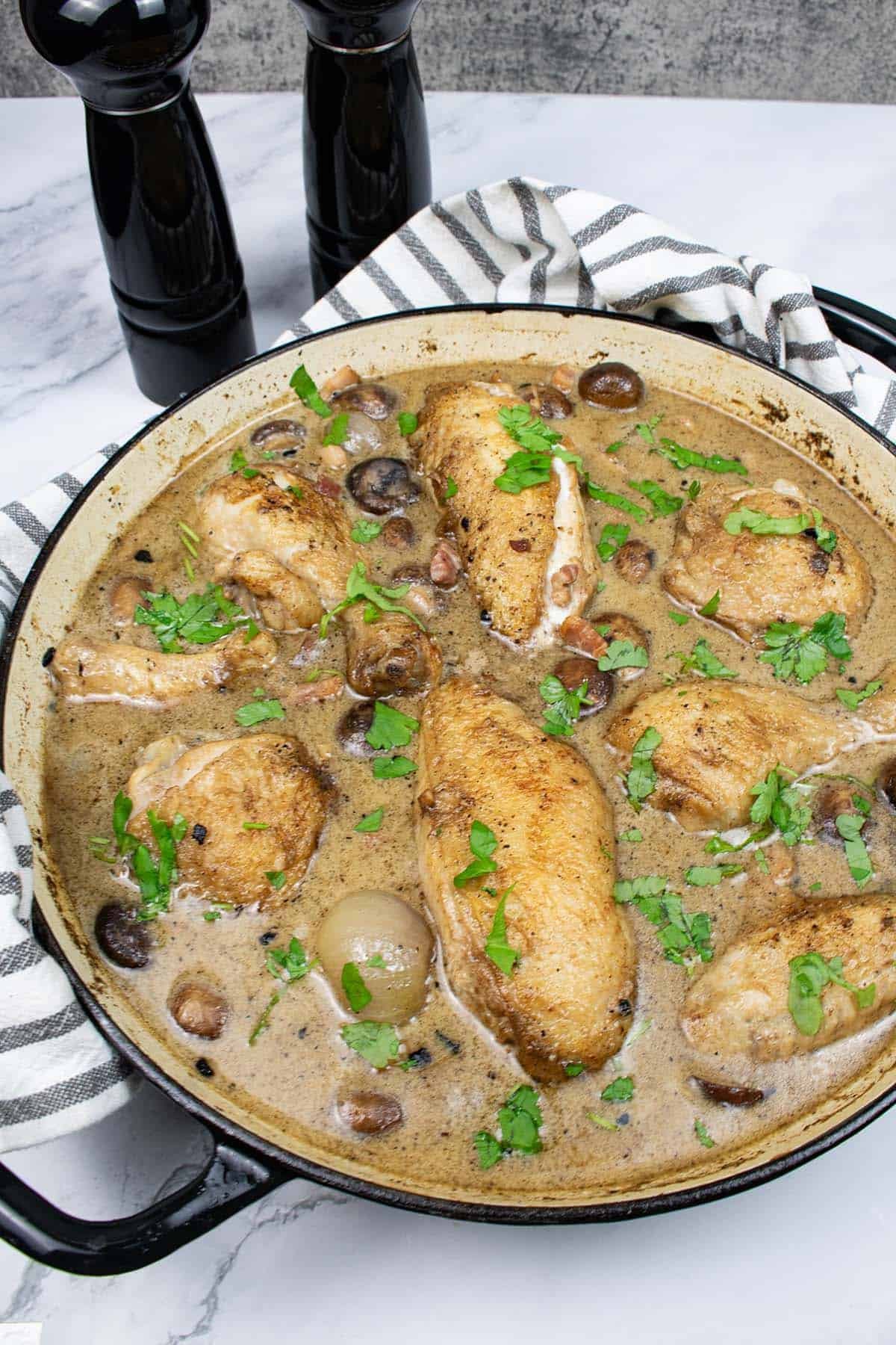 Coq au riesling in a round casserole with a black and white striped towel around the handles and black salt and pepper grinders in the background.