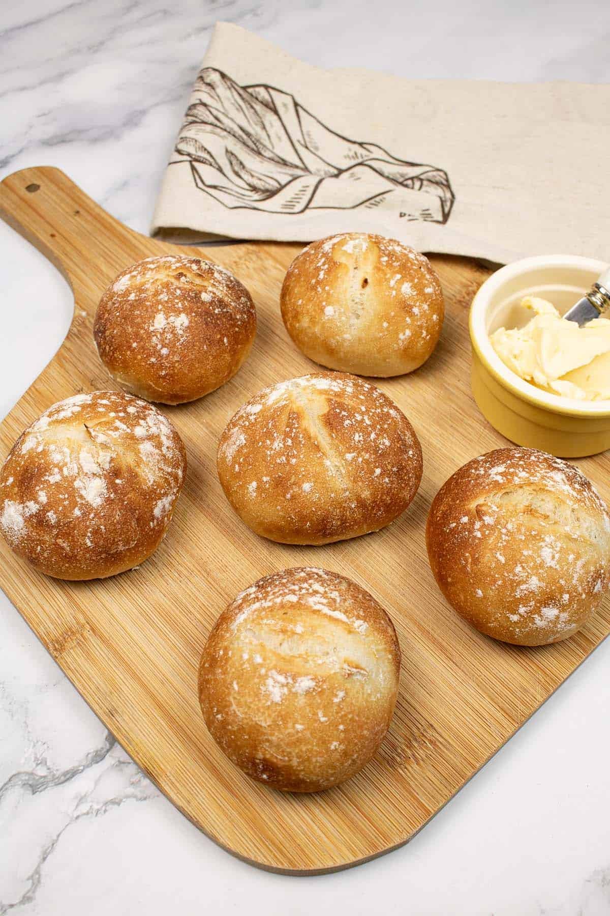 Crusty white dinner rolls on a board with a dish of butter with butter knife. Bread bag in the background.