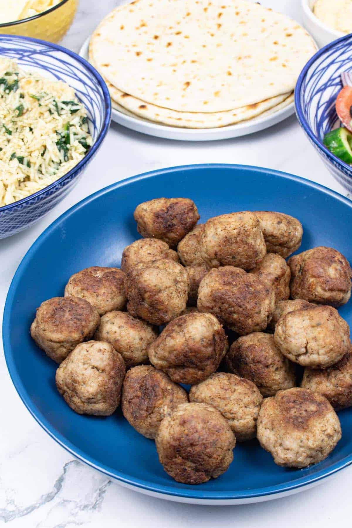 Greek lamb meatballs made in the Thermomix in a blue bowl with Greek rice with spinach, Greek salad and flatbreads on the side.