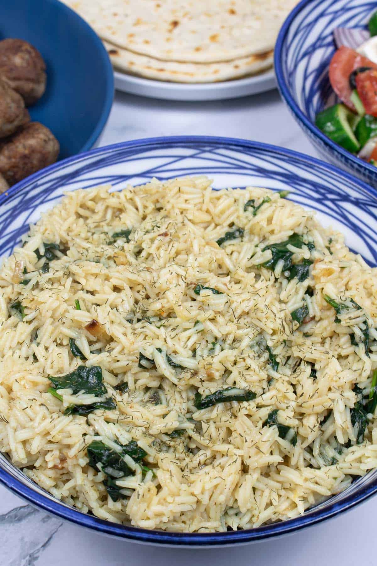 Greek rice with spinach in a blue and white patterned bowl, with bowls of lamb meatballs and Greek salad and a plate of flatbreads in the background.