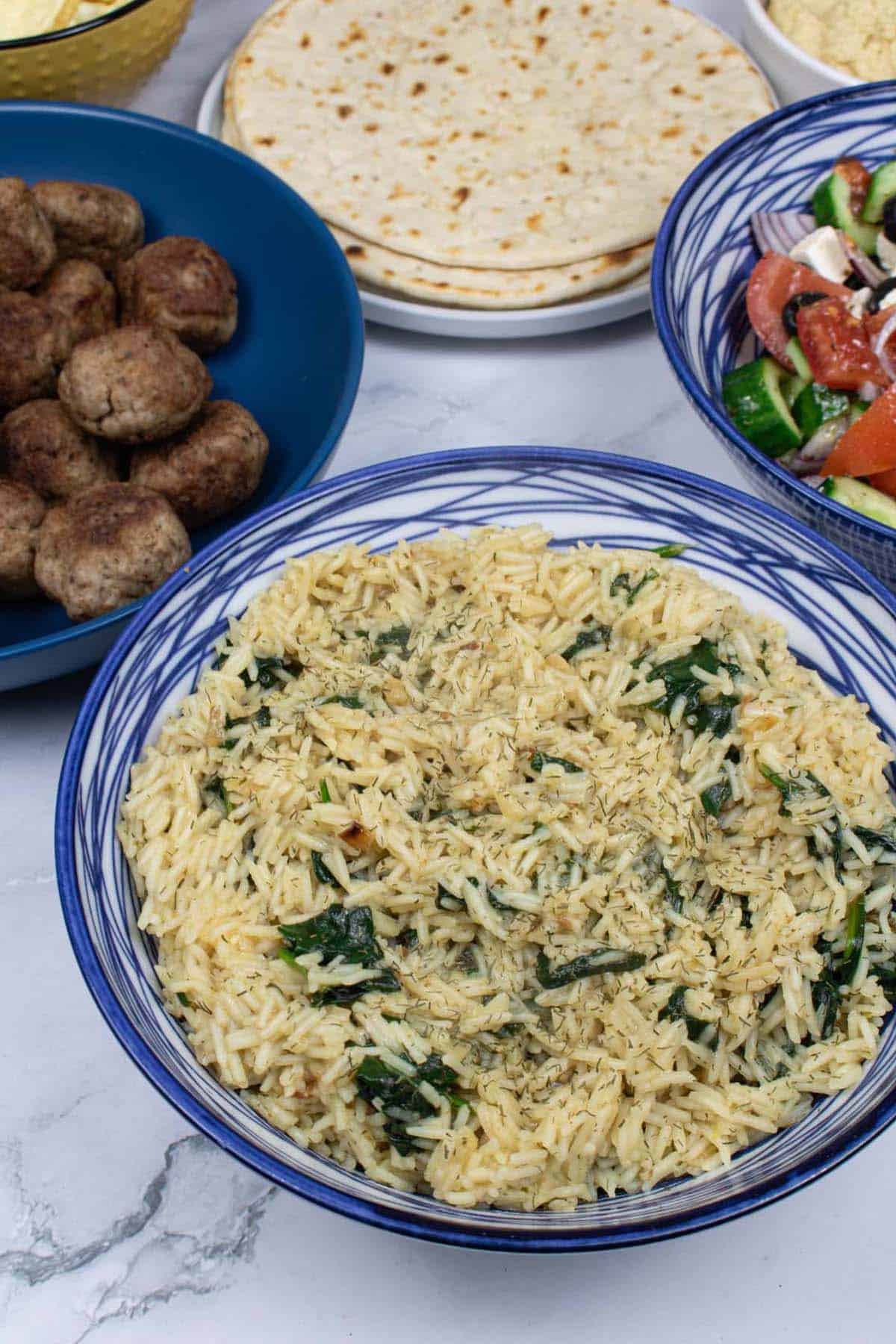 Greek rice with spinach in a blue and white patterned bowl, with bowls of lamb meatballs and Greek salad plus a plate of flatbreads in the background.