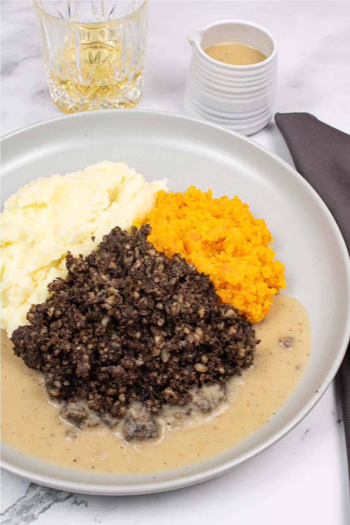 Haggis, neeps, tatties and whisky sauce on a grey dinner plate with a dram of whisky, small jug of whisky sauce and charcoal napkin on the side.