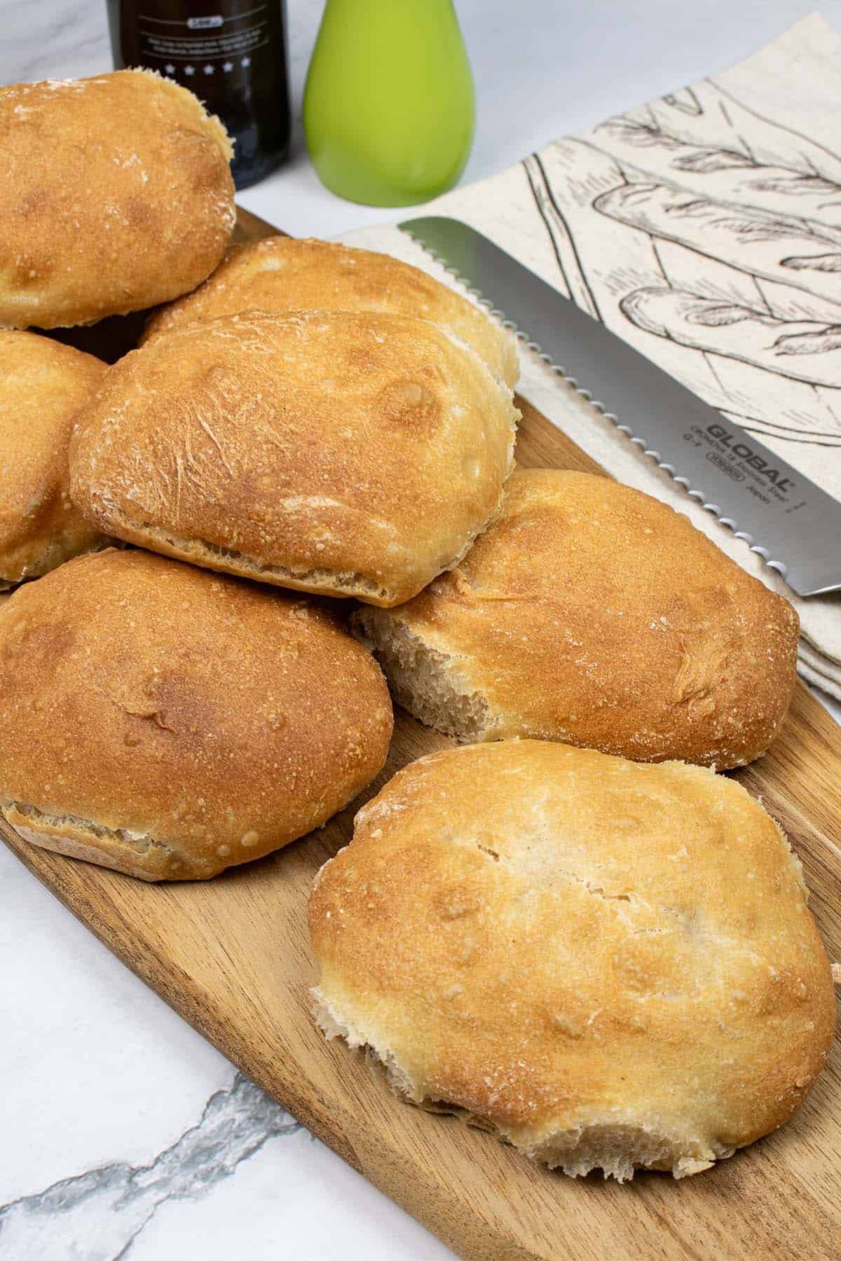 Homemade ciabatta bread rolls on a board with a bread knife, bread bag, oil and pepper on the side.