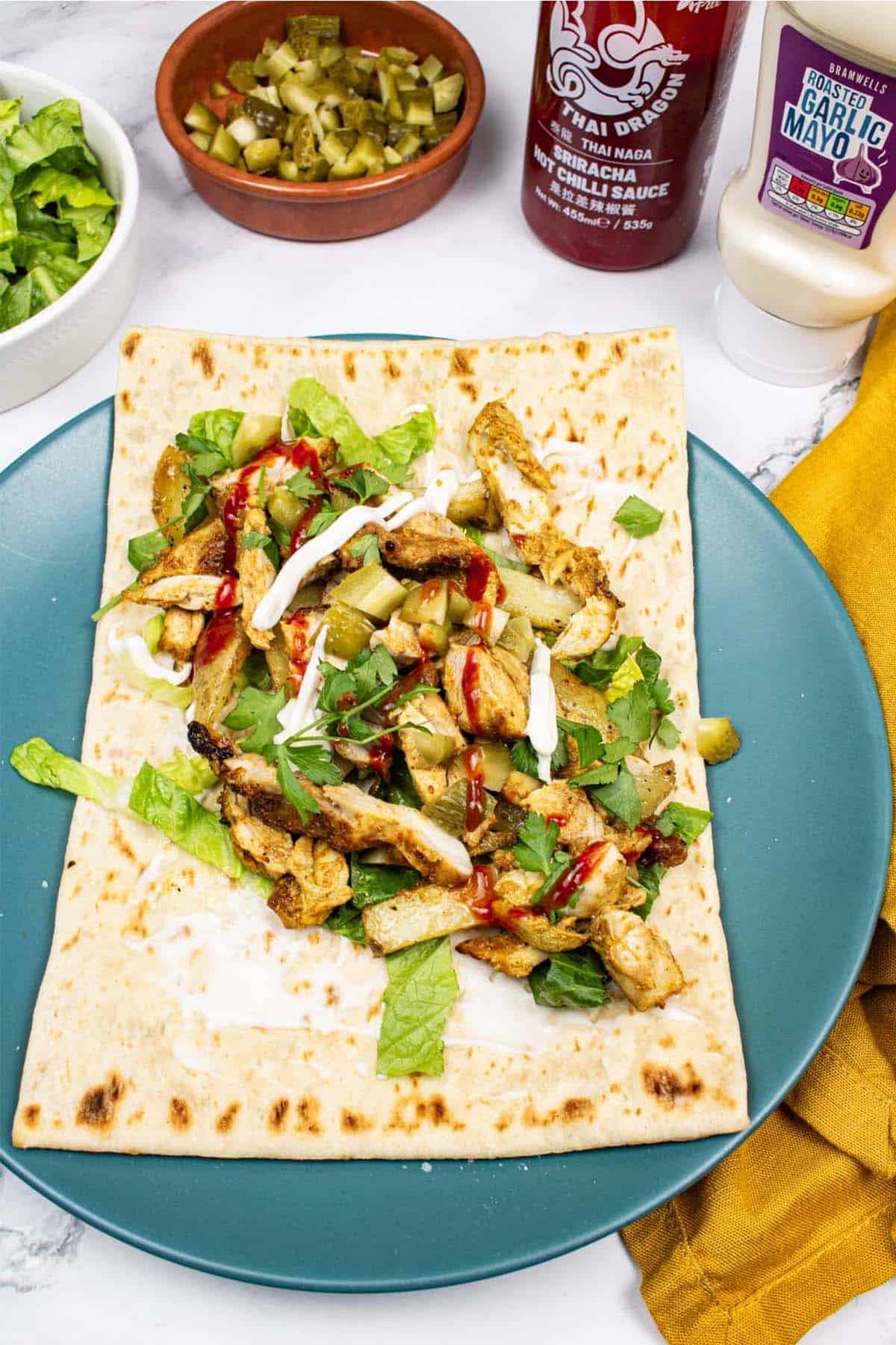 Kebab king shawarma assembled on a teal dinner plate with dishes of shredded lettuce, chopped gherkins and bottles of sriracha and garlic mayo on the side.