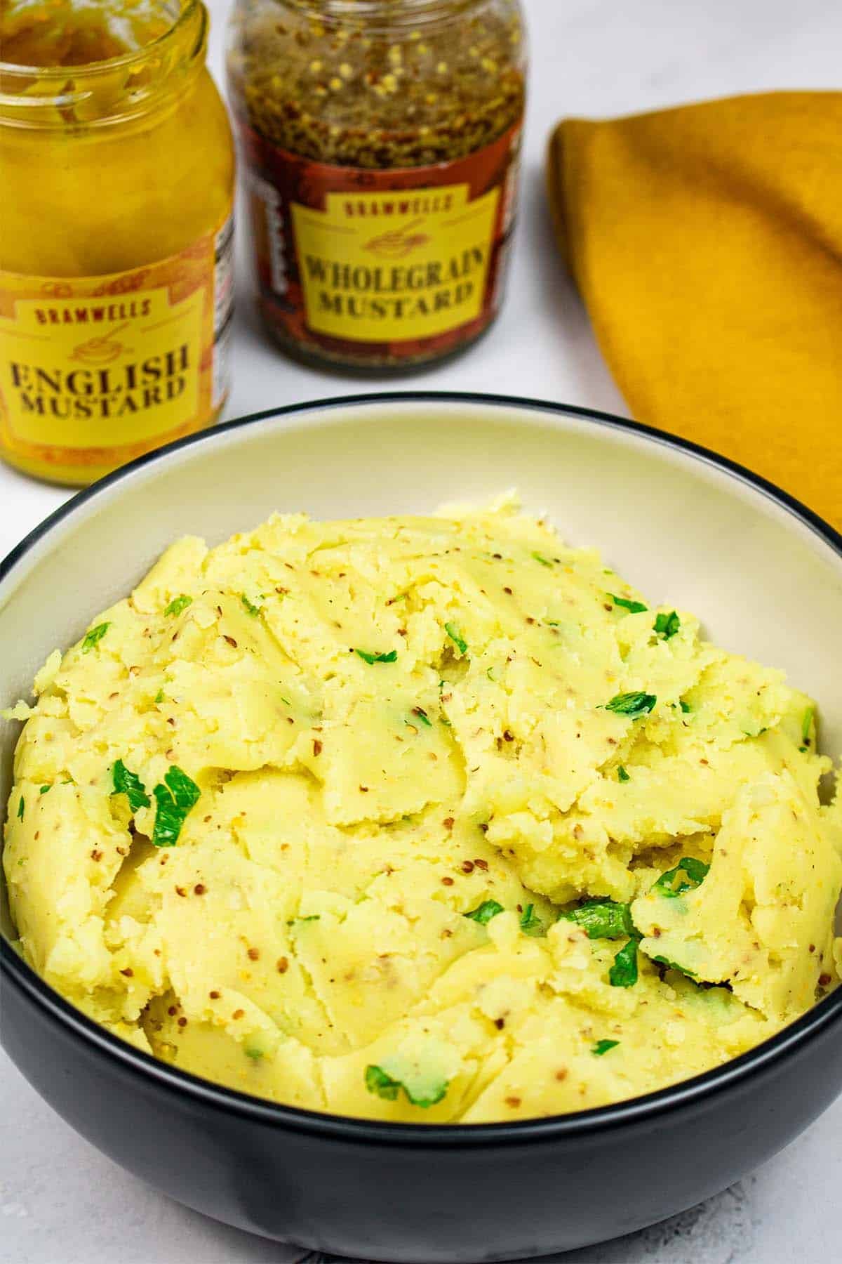 Mustard mash in a black and cream bowl with jars of English and wholegrain mustards, and mustard napkin on the side