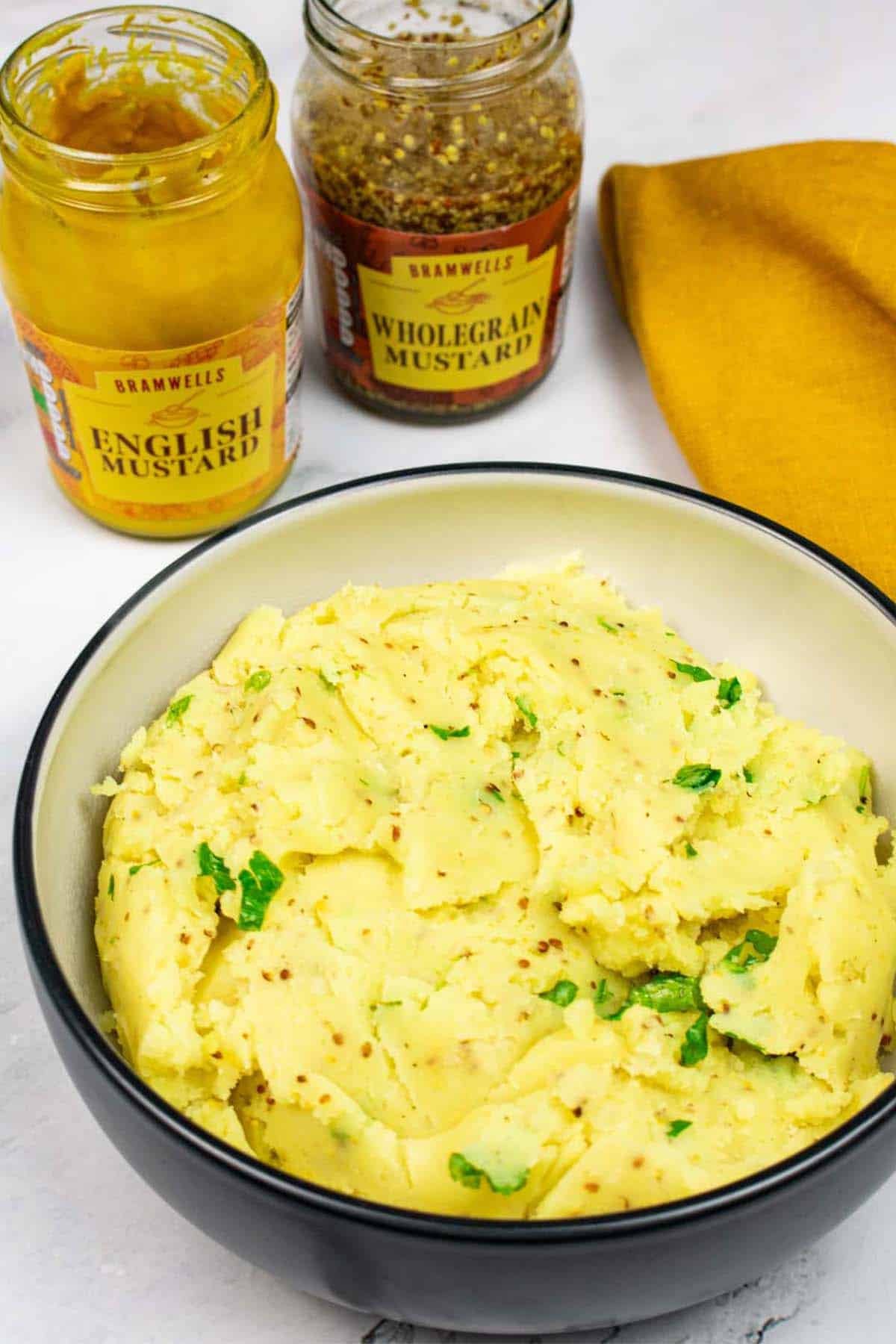 Mustard mash in a black and cream bowl with jars of English and wholegrain mustards, and mustard napkin on the side