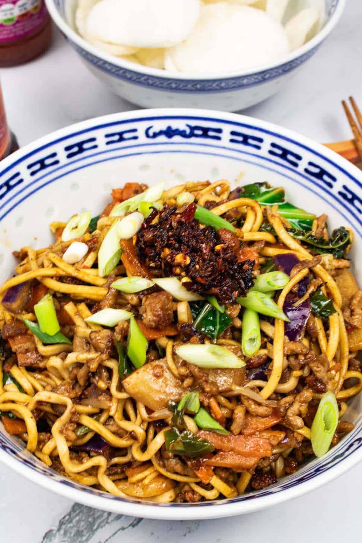 Pork noodle stir-fry in and oriental bowl with a bowl of prawn crackers, jar of crispy chilli in oil, bottle of sweet chilli sauce and chopsticks on the side.