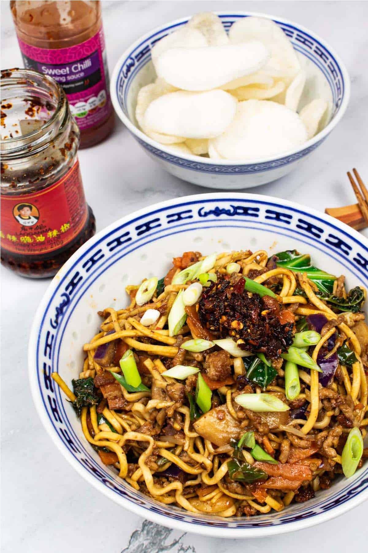 Pork noodle stir-fry in and oriental bowl with a bowl of prawn crackers, jar of crispy chilli in oil, bottle of sweet chilli sauce and chopsticks on the side.