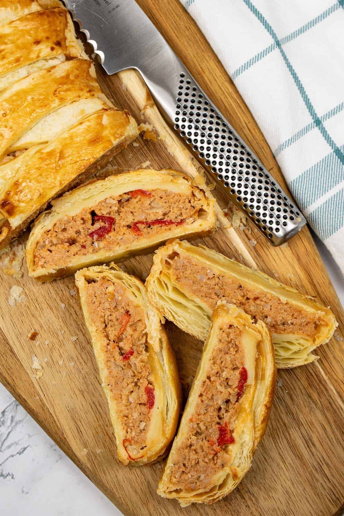 Spanish-flavoured sausagemeat plait made in Thermomix on a wooden board with 4 pieces sliced, a bread knife and teal and white checked towel on the side.