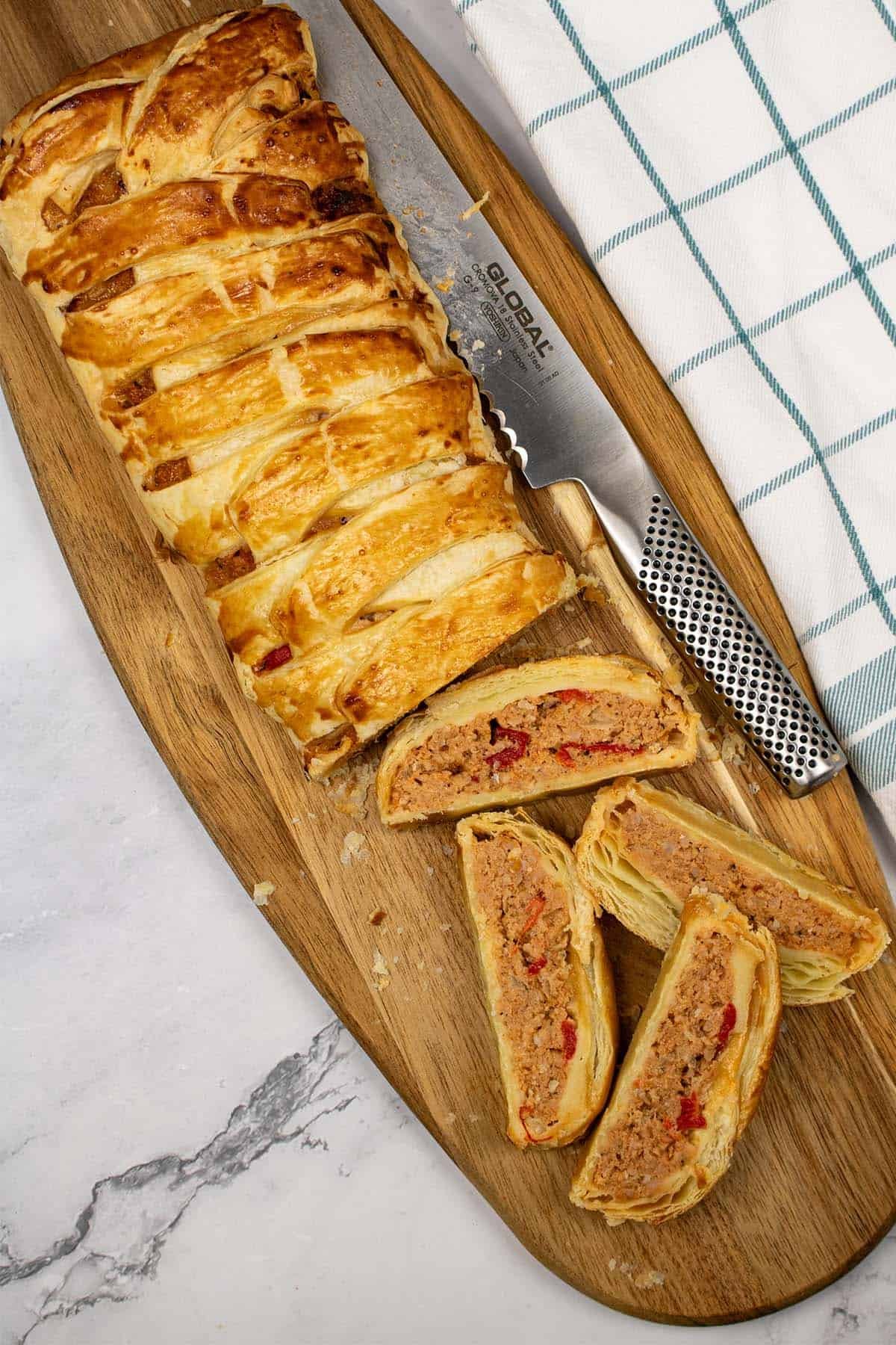 Spanish-flavoured sausagemeat plait made in Thermomix on a wooden board with 4 pieces sliced, a bread knife and teal and white checked towel on the side.