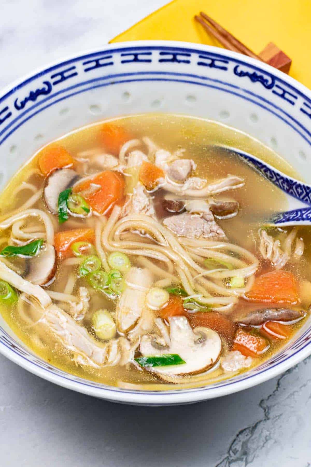 Chinese chicken noodle soup in a blue and white oriental bowl with spoon. A yellow napkin and chopsticks on the side.