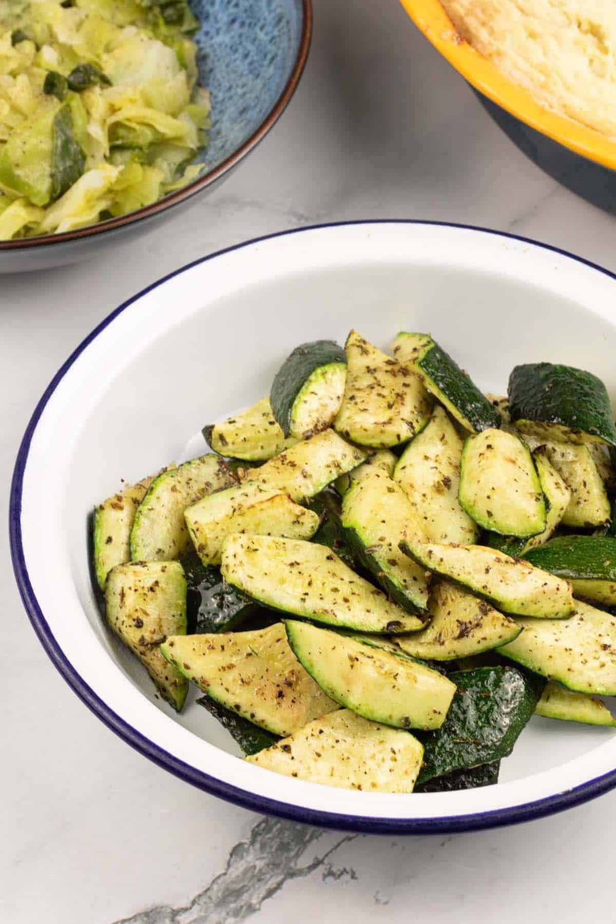 Easy air fryer courgettes in a round blue rimmed white enamel dish with a bowl of cabbage and a pie in the background.