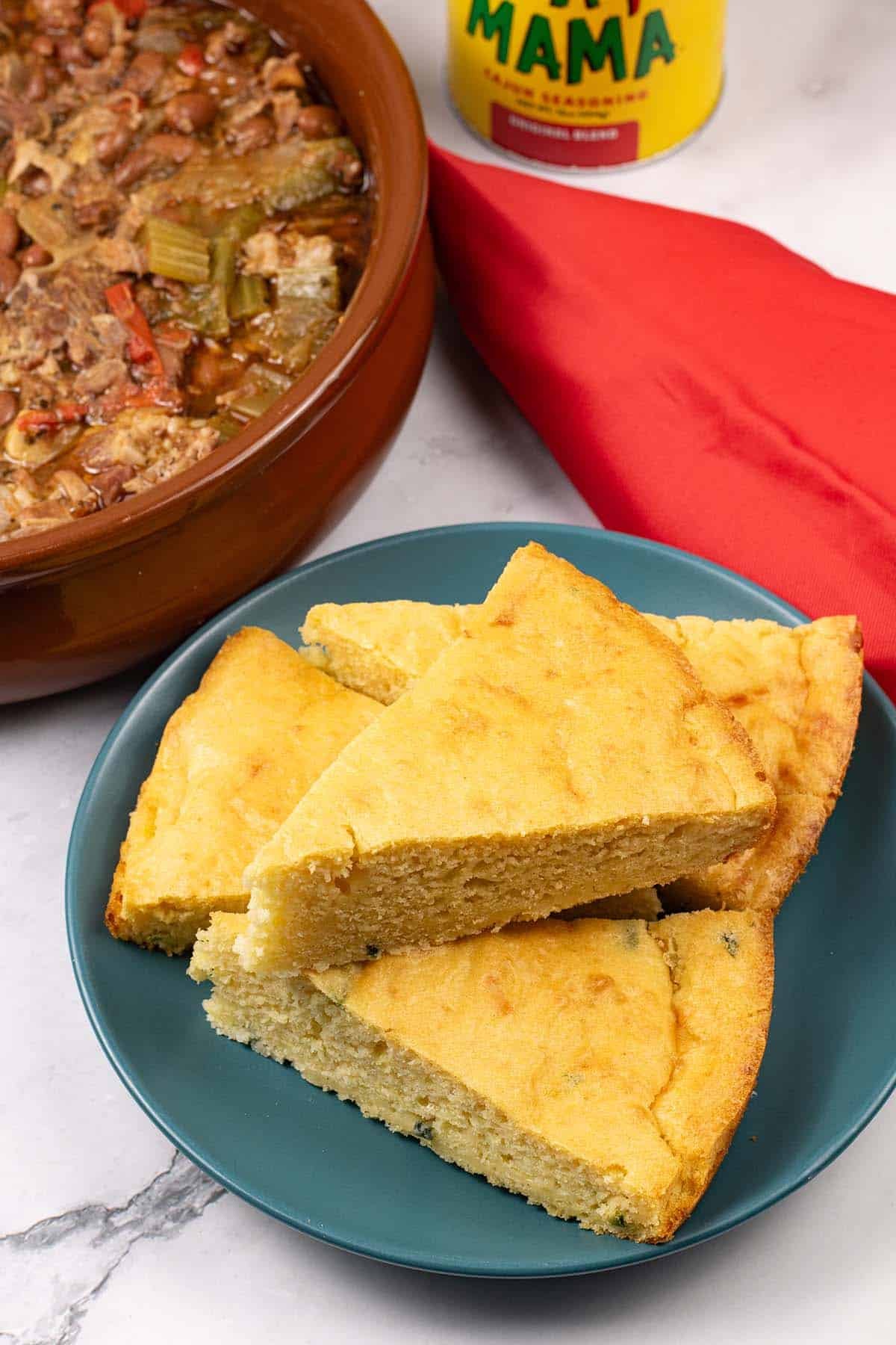 Slices of easy homemade cornbread on a teal side plate with a dish of cajun pork, tub of cajun seasoning and red napkin on the side.