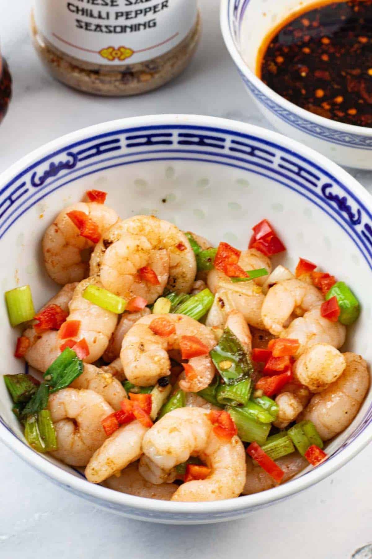 Easy salt and chilli pepper prawns in a blue and white oriental bowl, with a bowl of dipping sauce and a jar of Chinese seasoning in the background.