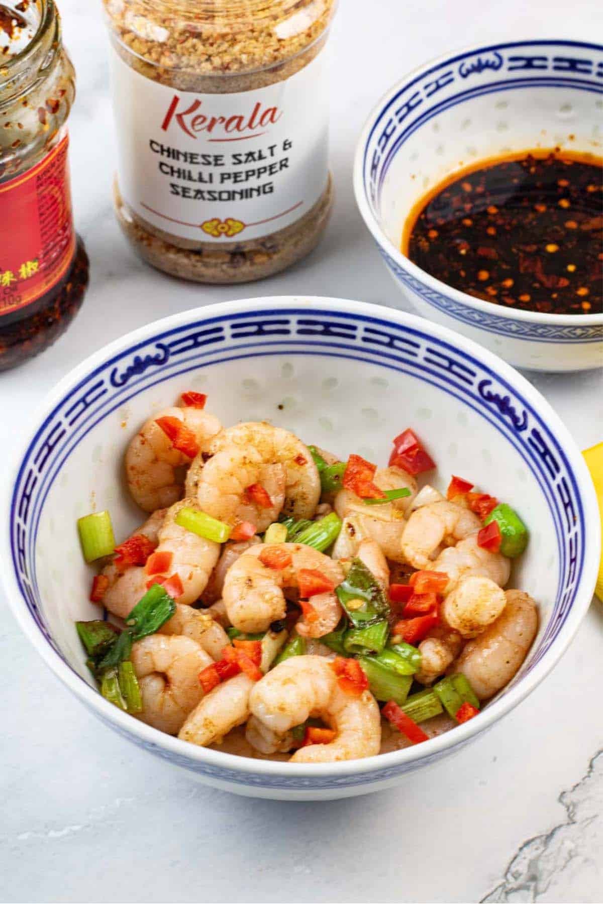 Easy salt and chilli pepper prawns in a blue and white oriental bowl, with a bowl of dipping sauce and jars of crispy chilli in oil and Chinese seasoning in the background.