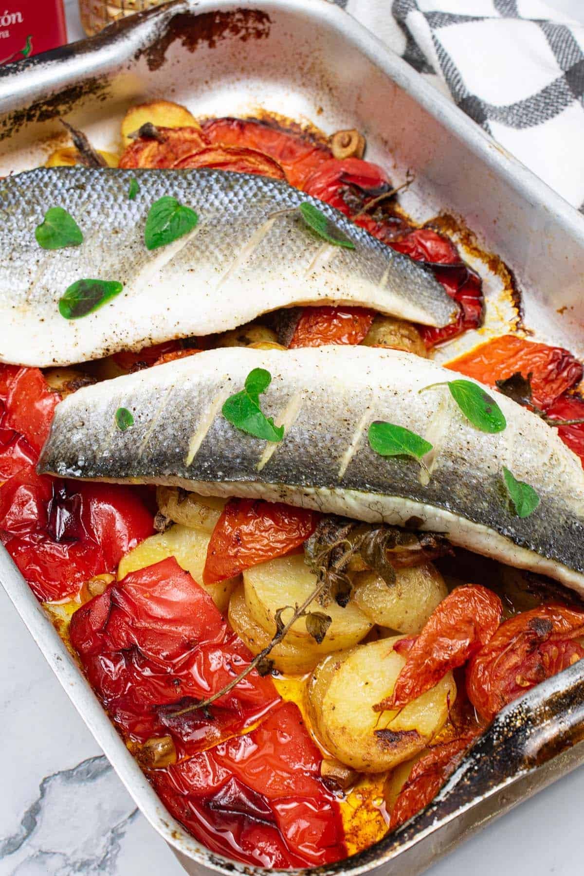 Two portions of easy sea bass with roasted peppers and potatoes in a roasting tin. A white checked towel on the side.