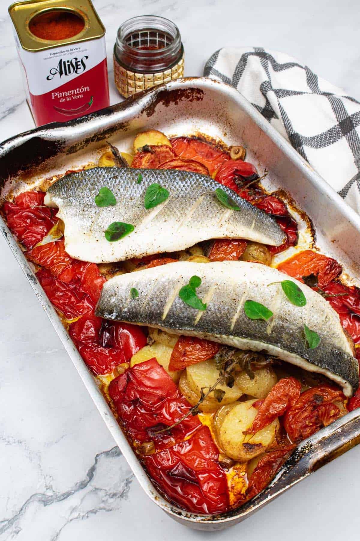 Two portions of easy sea bass with roasted peppers and potatoes in a roasting tin. A tin of pimenton, a jar of saffron and a black and white checked towel on the side.