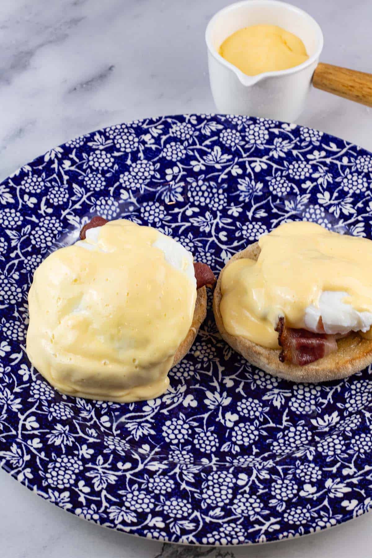 Eggs benedict with quick hollandaise sauce on a blue and white patterned dinner plate, with a small jug of sauce on the side.