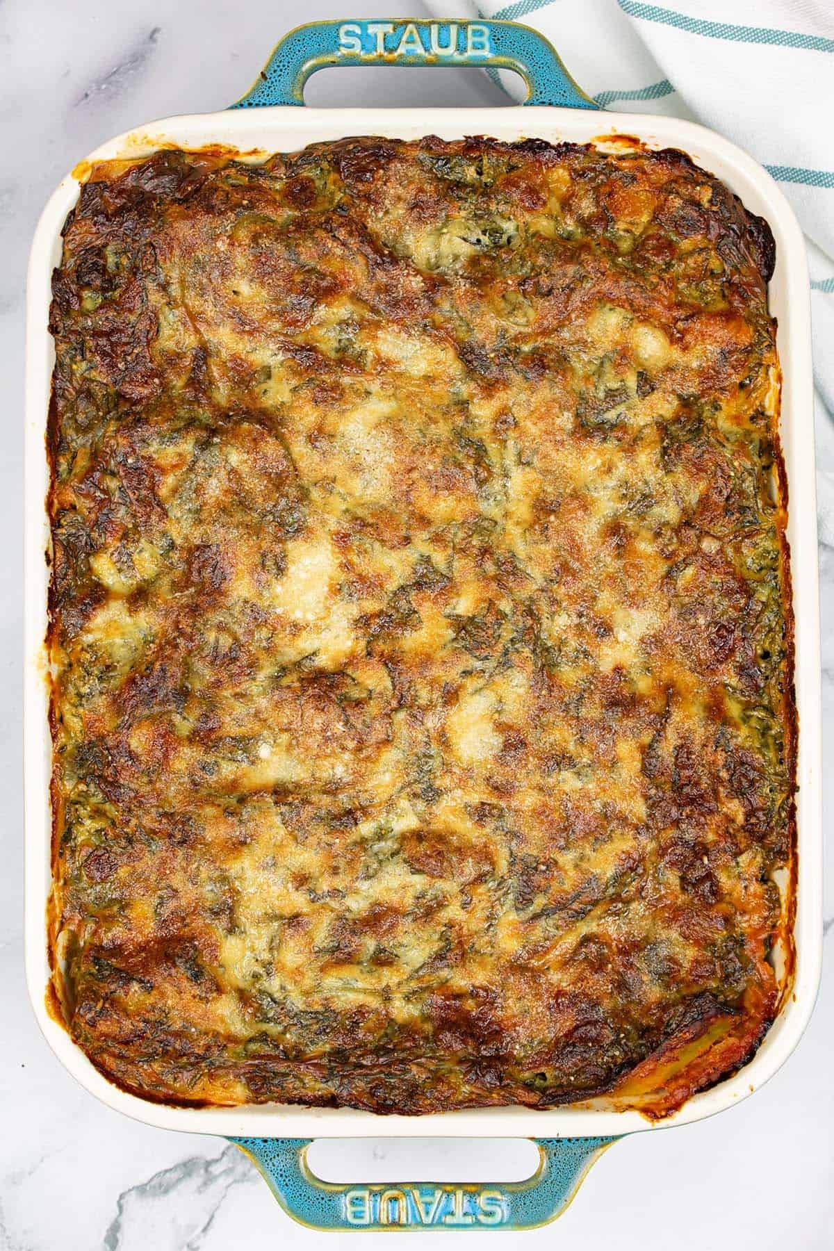 Healthy turkey lasagne in a rectangular Staub dish with a teal and white striped towel on the side.