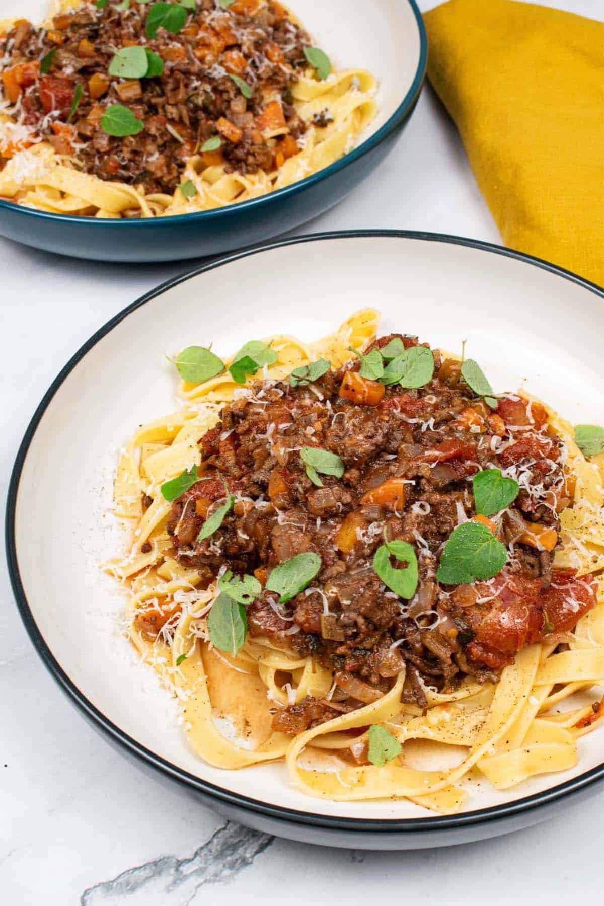 2 pasta bowls of lamb, tomato and oregano ragu with tagliatelle and a mustard napkin on the side.