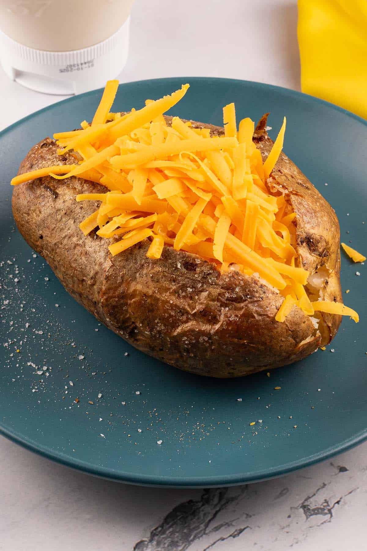 Perfect air fryer baked potato on a teal side plate topped with grated cheese. A yellow napkin and bottle of mayo on the side.