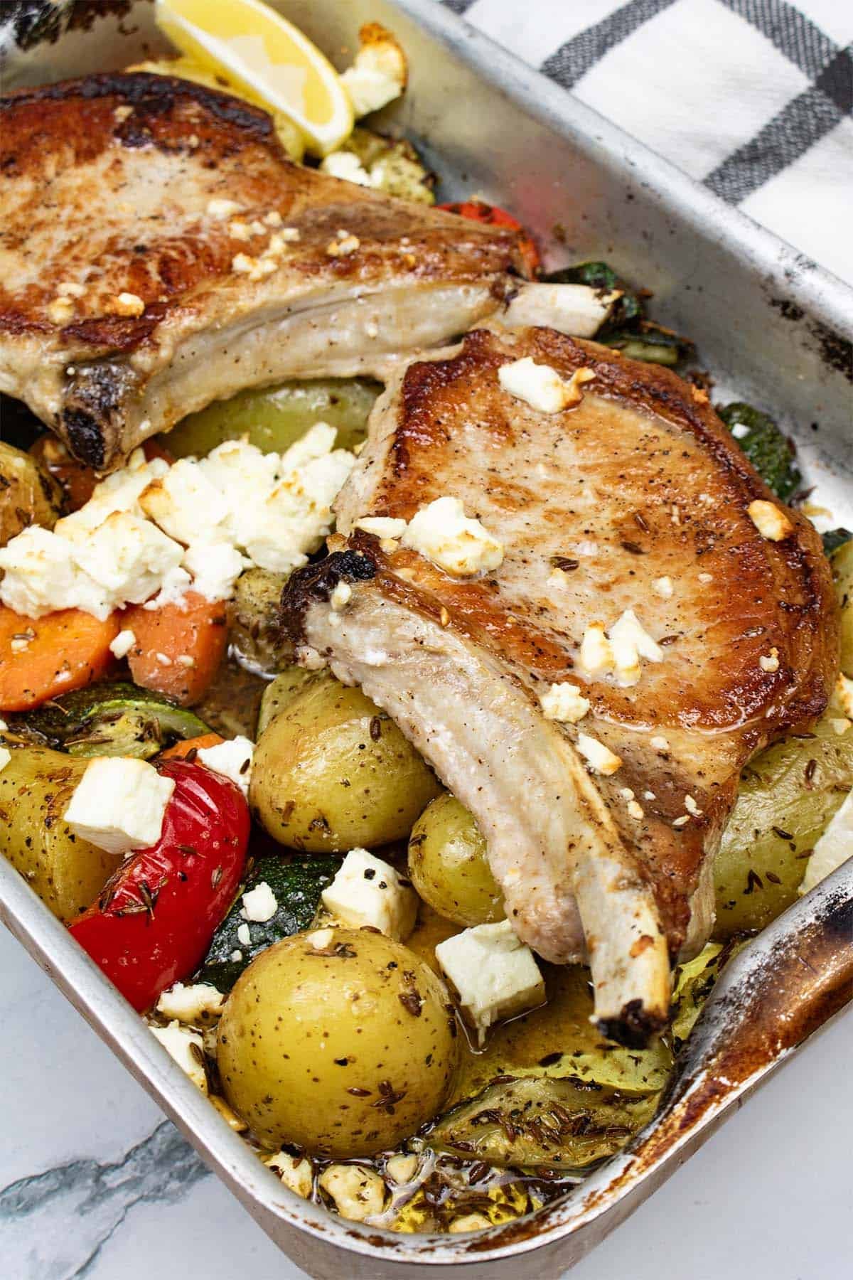 Pork chops with roast vegetables and feta in a roasting tin with a black and white checked towel on the side.