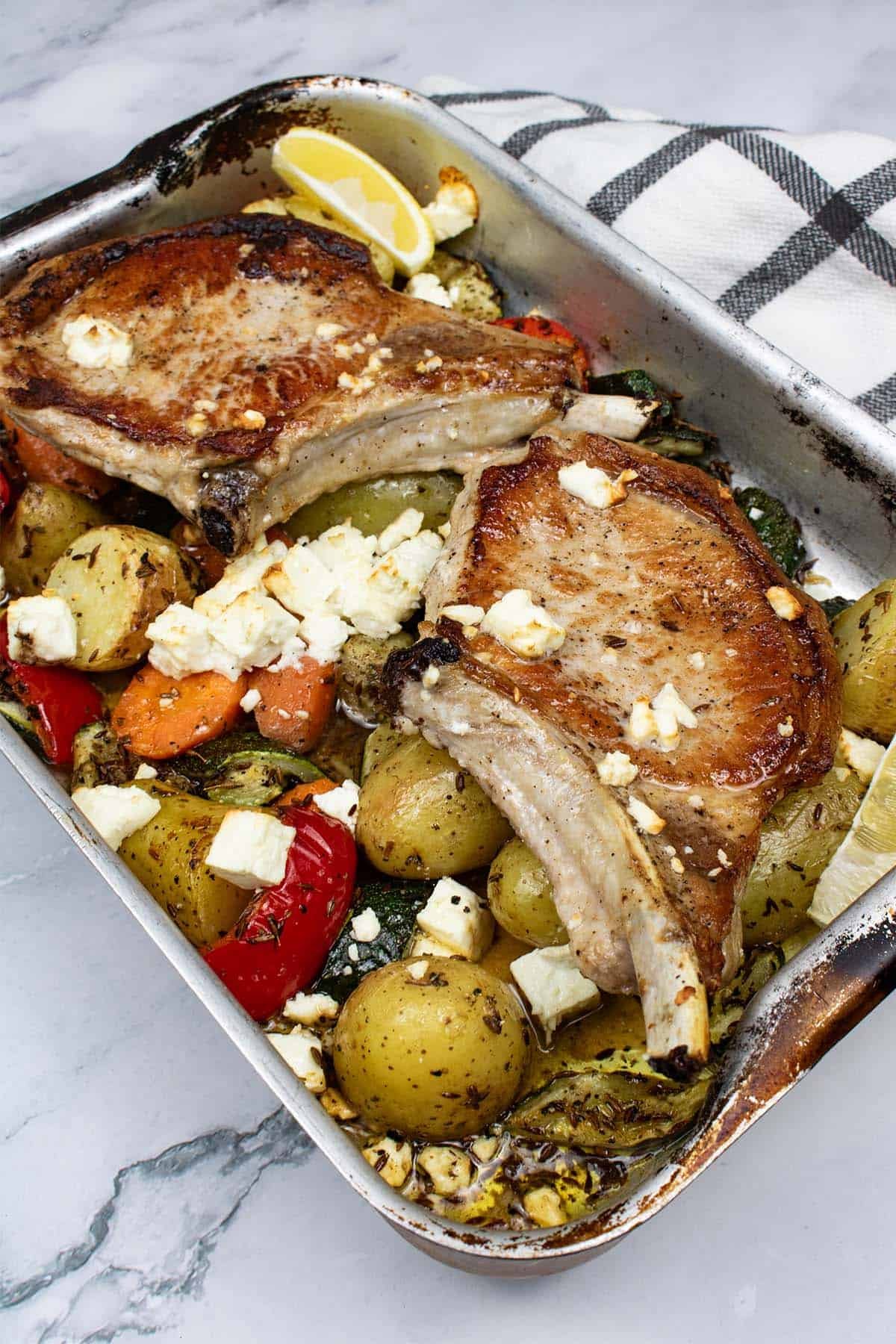 Pork chops with roast vegetables and feta in a roasting tin with a black and white checked towel on the side.