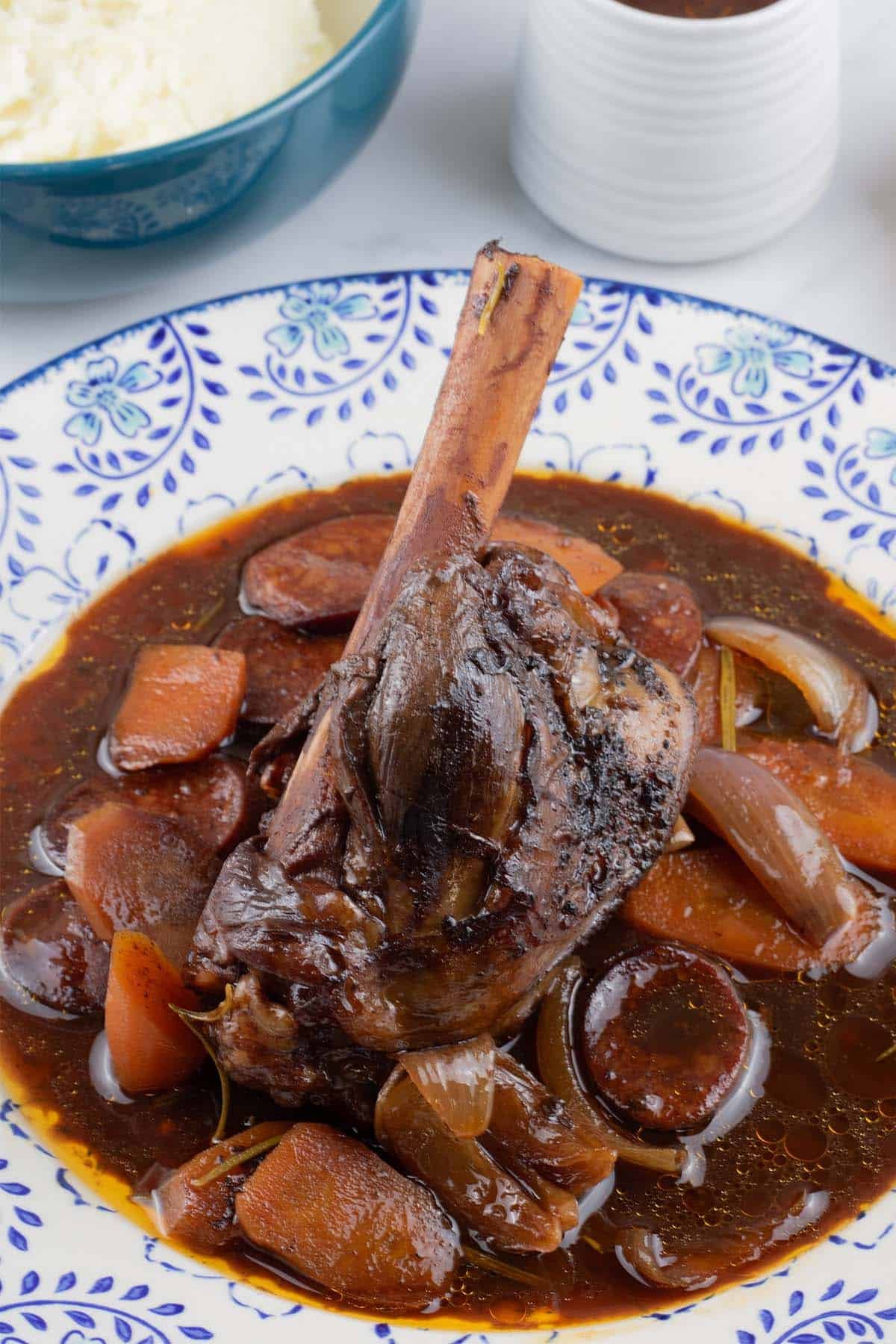 Rioja-braised lamb shank with chorizo and garlic on a blue and white patterned dinner plate, with a bowl of mashed potatoes and small jug of sauce on the side.