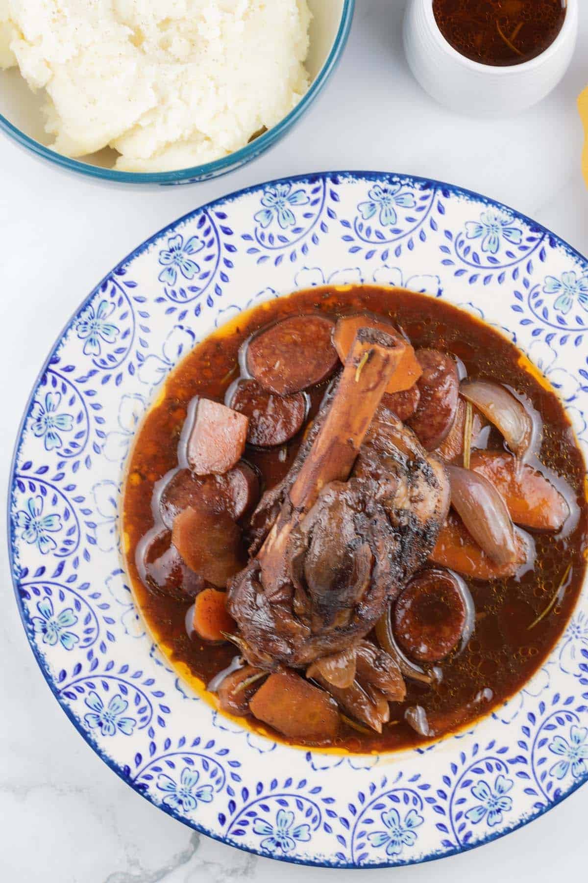 Rioja-braised lamb shank with chorizo and garlic on a blue and white patterned dinner plate, with a bowl of mashed potatoes and small jug of sauce on the side.