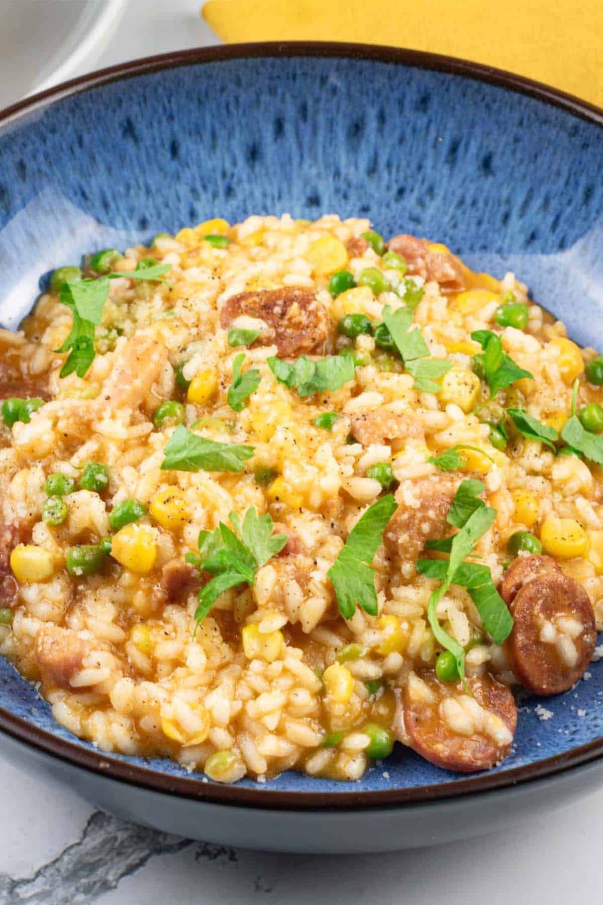 A close up portion of sausage and pea risotto made in the Thermomix in a blue patterned bowl with a mustard napkin on the side.