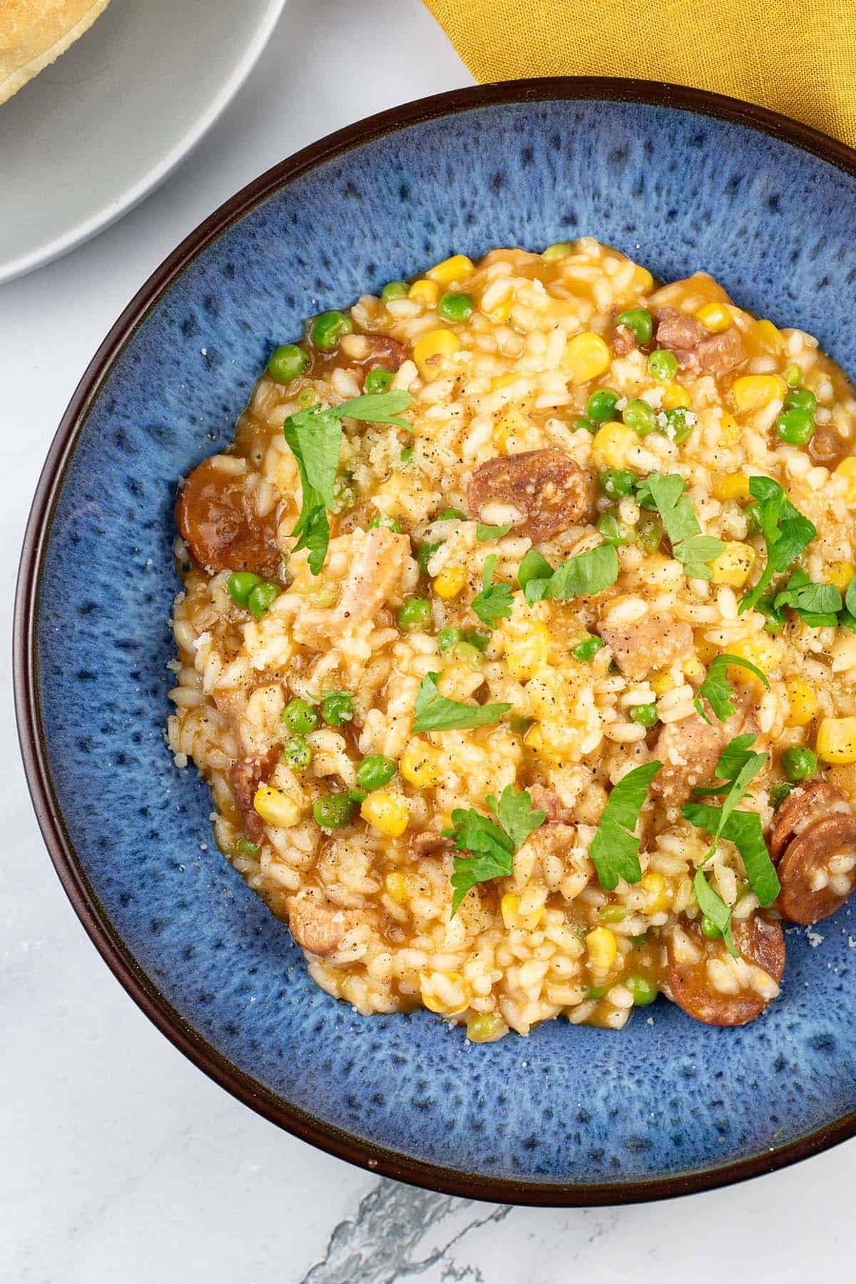 A portion of sausage and pea risotto made in the Thermomix in a blue patterned bowl with a crusty bread roll and mustard napkin on the side.