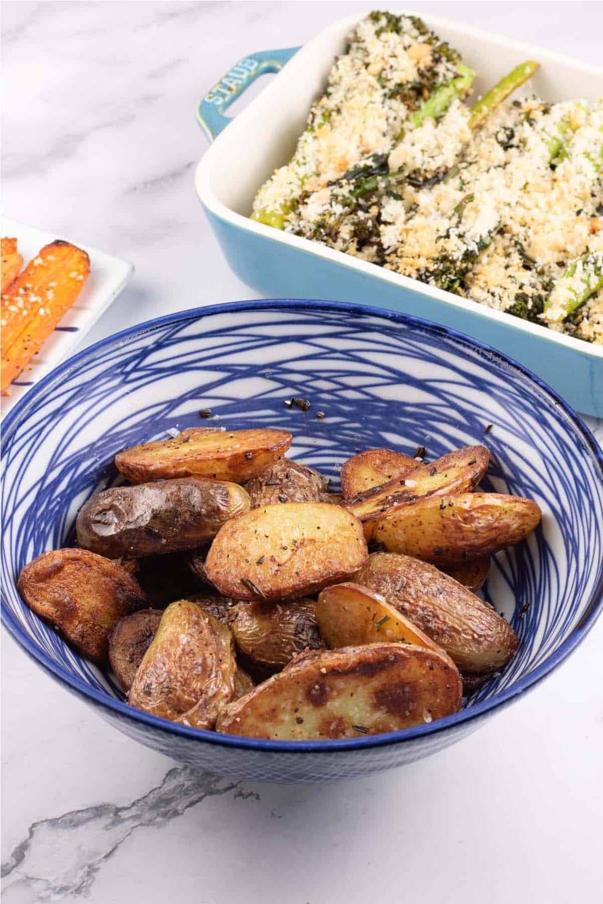 Sautéed new potatoes with rosemary in a blue and white striped bowl, with dishes of za'atar carrots and tenderstem broccoli on the side.