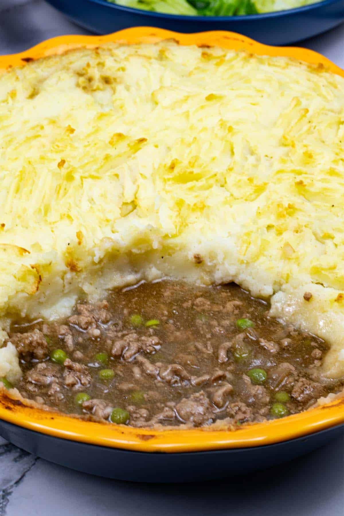 Shepherd's pie in a round yellow and blue pie dish with a portion of potatoes removed, and a bowl of buttered cabbage in the background.