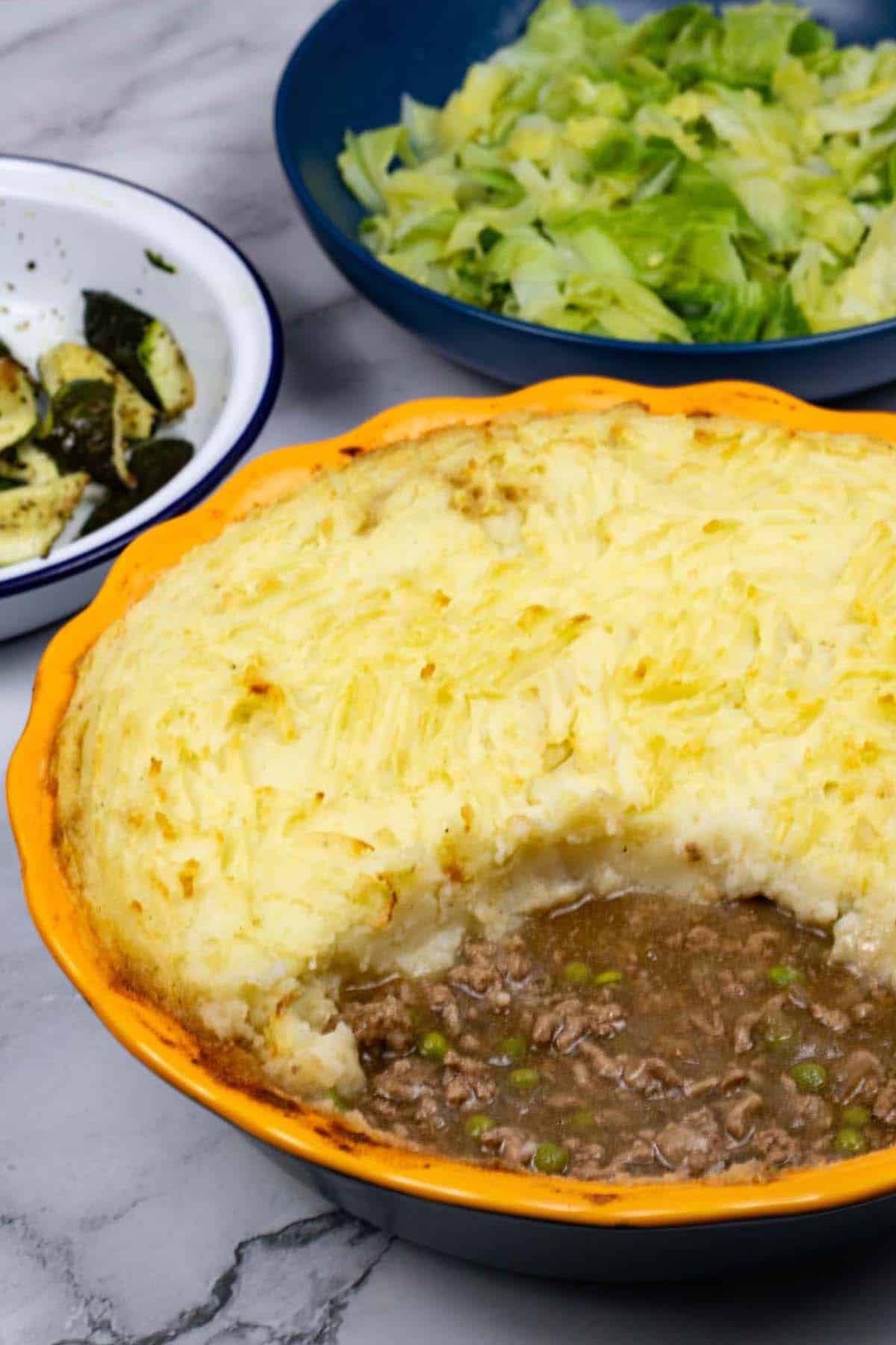 Shepherd's pie in a round yellow and blue pie dish with a portion of potatoes removed, and a bowl of buttered cabbage in the background.