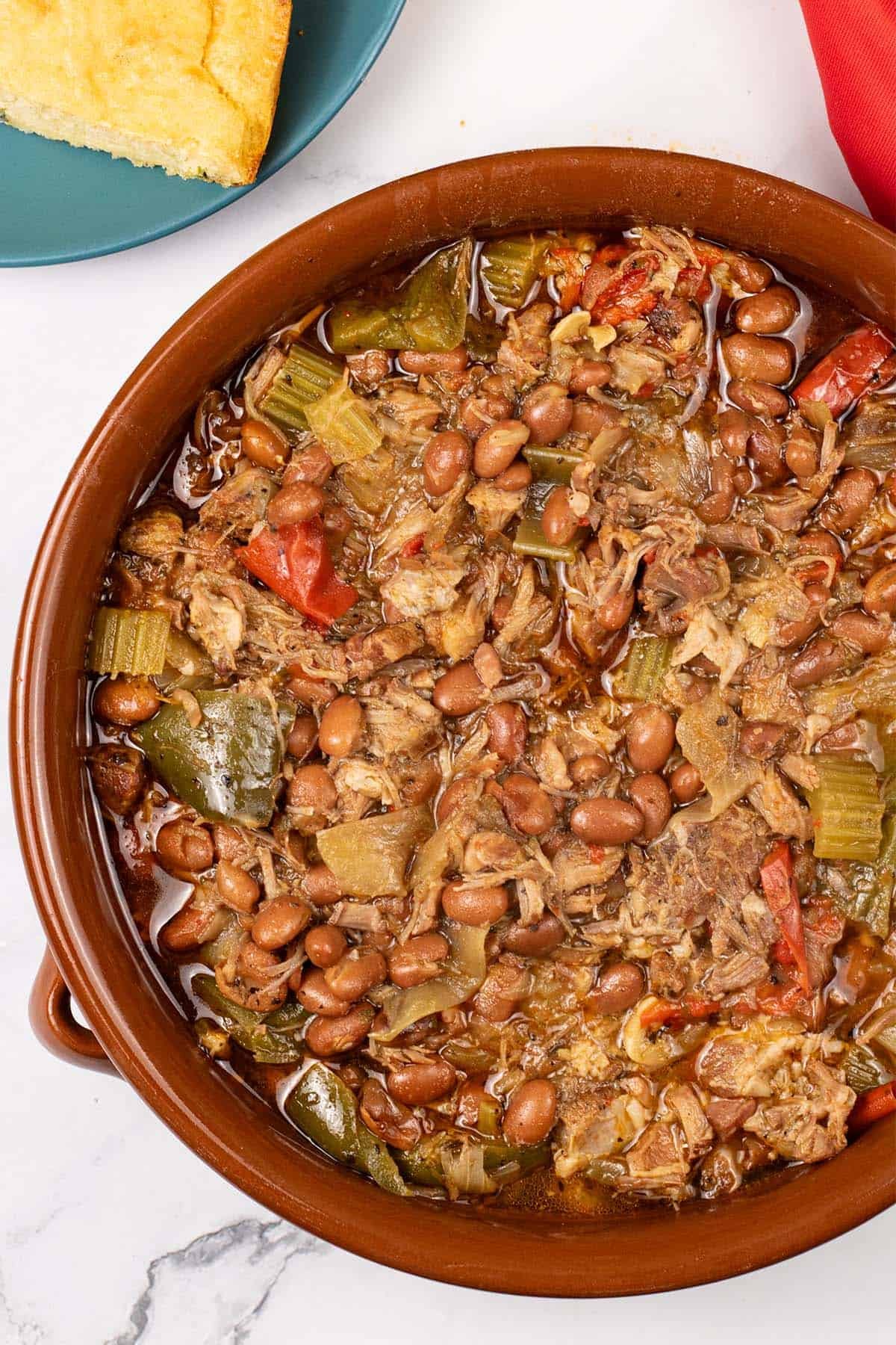 Slow cooker cajun pork and red beans in a large round terracotta dish with a plate of cornbread and red napkin on the side.