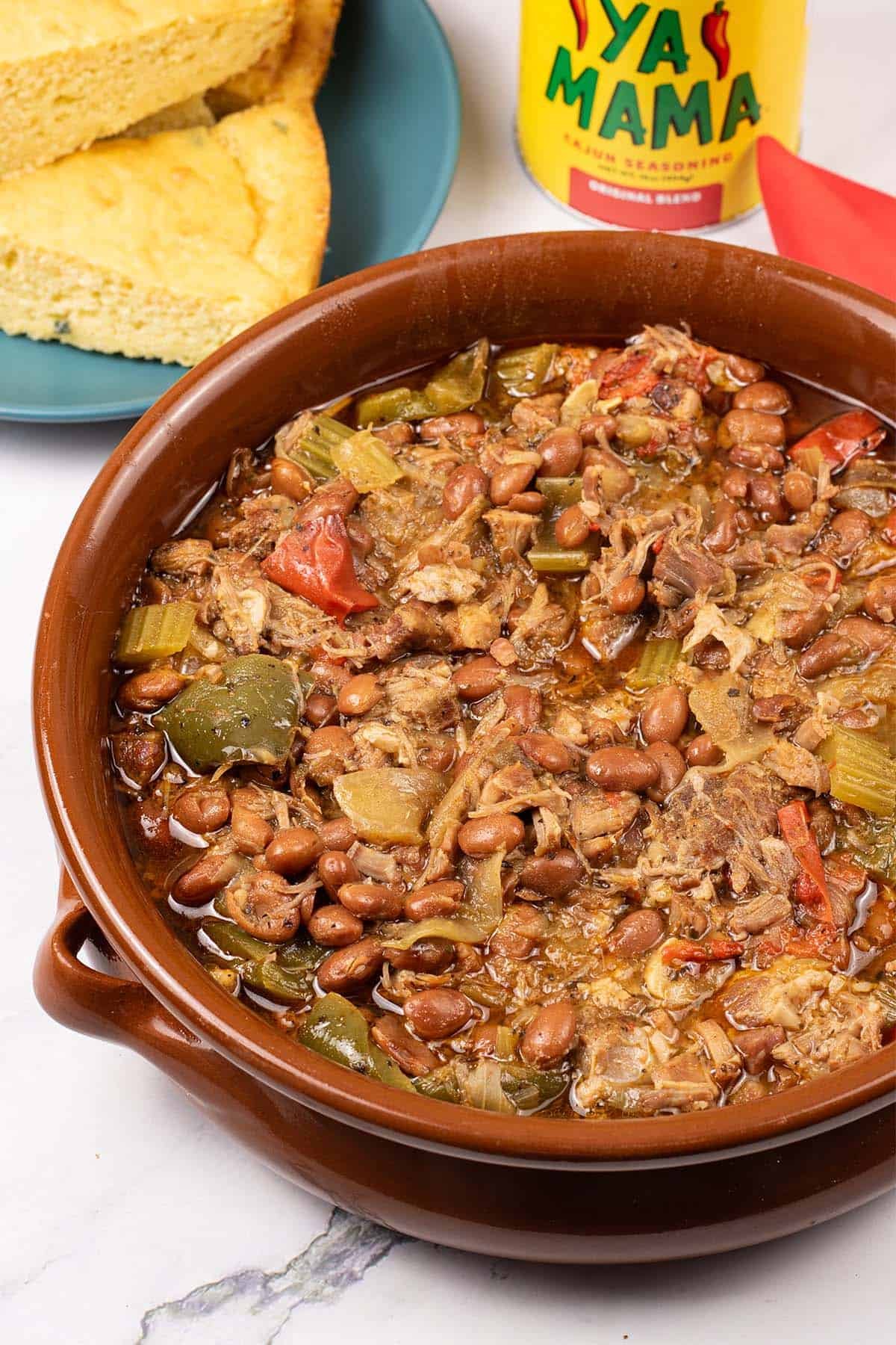 Slow cooker cajun pork and red beans in a large round terracotta dish with a plate of cornbread, tub of cajun seasoning and red napkin on the side.