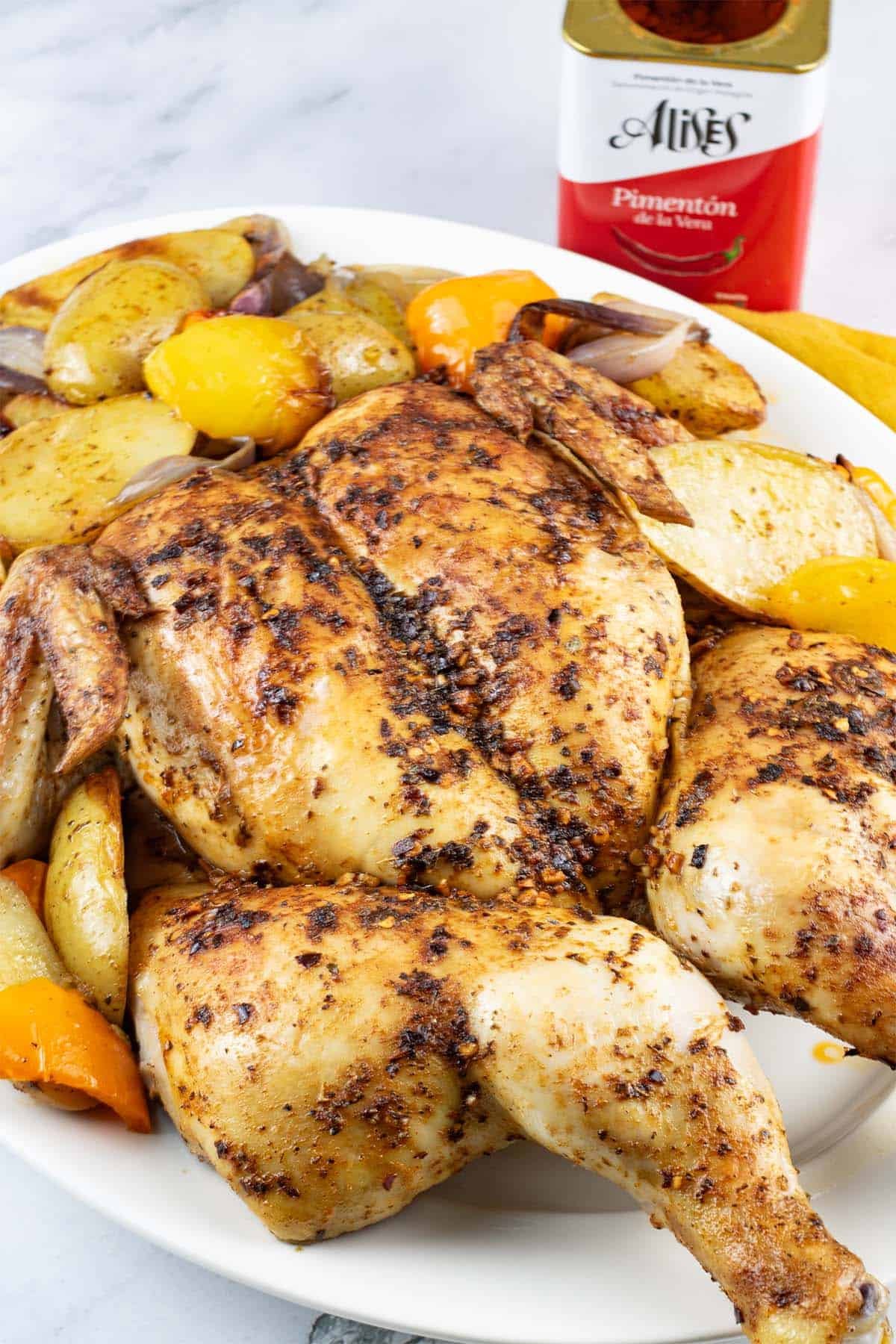 Spanish spatchcock chicken with vegetables on a white platter with tin of hot smoked pimenton and a mustard napkin on the side.